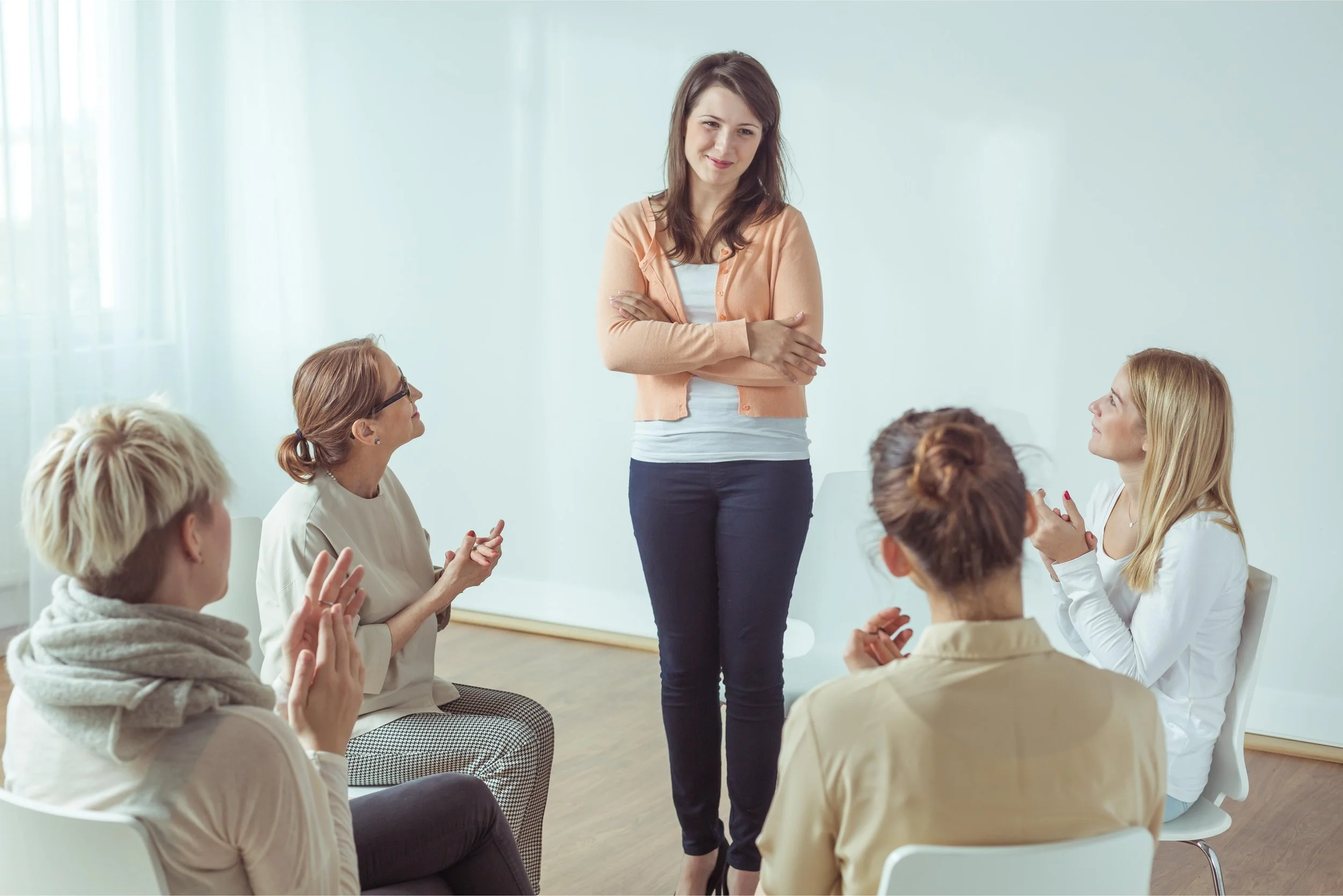 A woman standing with arms crossed in front of a group of seated women in a bright, white room, leading a discussion or presentation.
