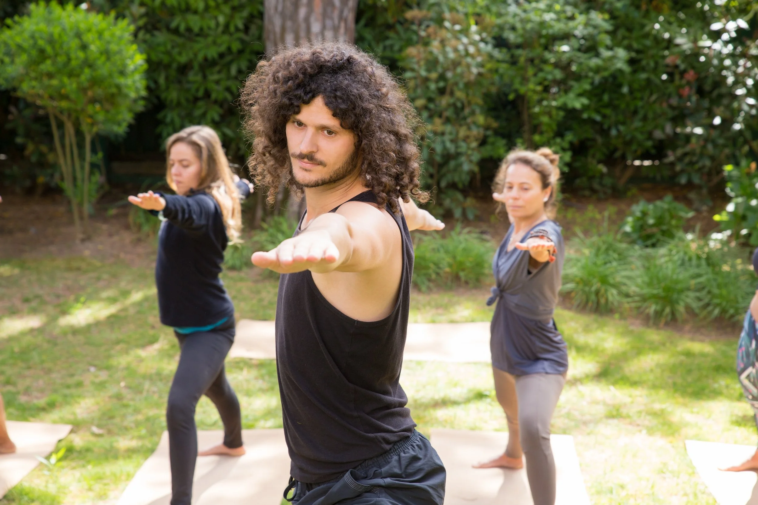 A man with curly hair in a black tank top practicing yoga outdoors with three women in the background also doing yoga, surrounded by trees and greenery.