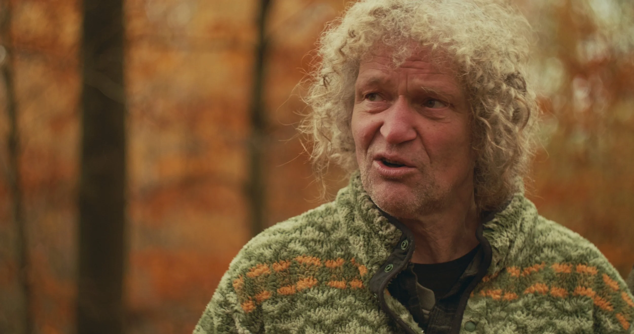 Ein Mann mit lockigem grauem Haar steht in einem Wald mit Herbstlaub. Er trägt eine grün-braune Jacke und schaut nach rechts.