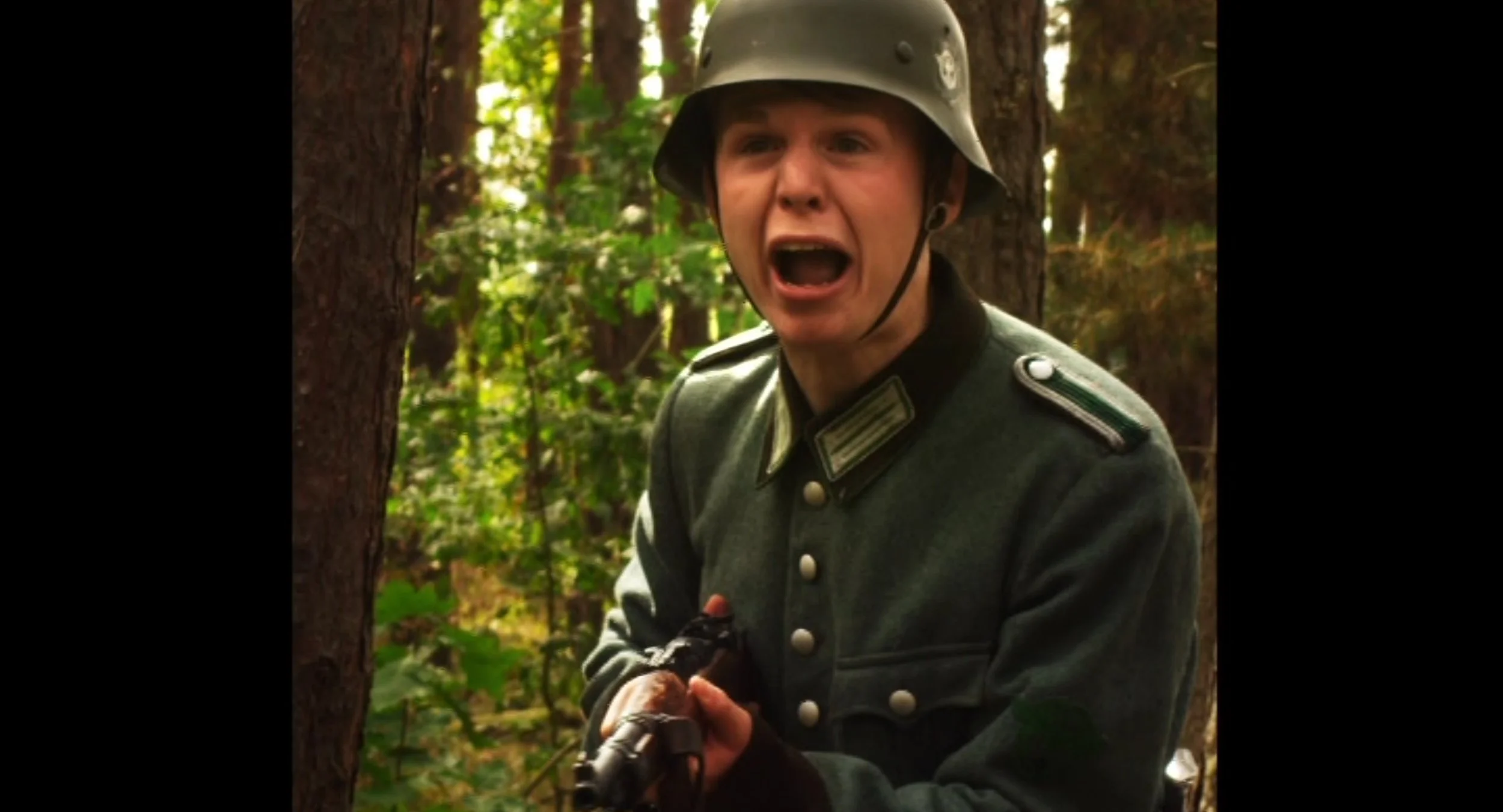 Ein Mann in einer deutschen Uniform und Helm schimpft in einem Wald, hält eine Waffe in den Händen.