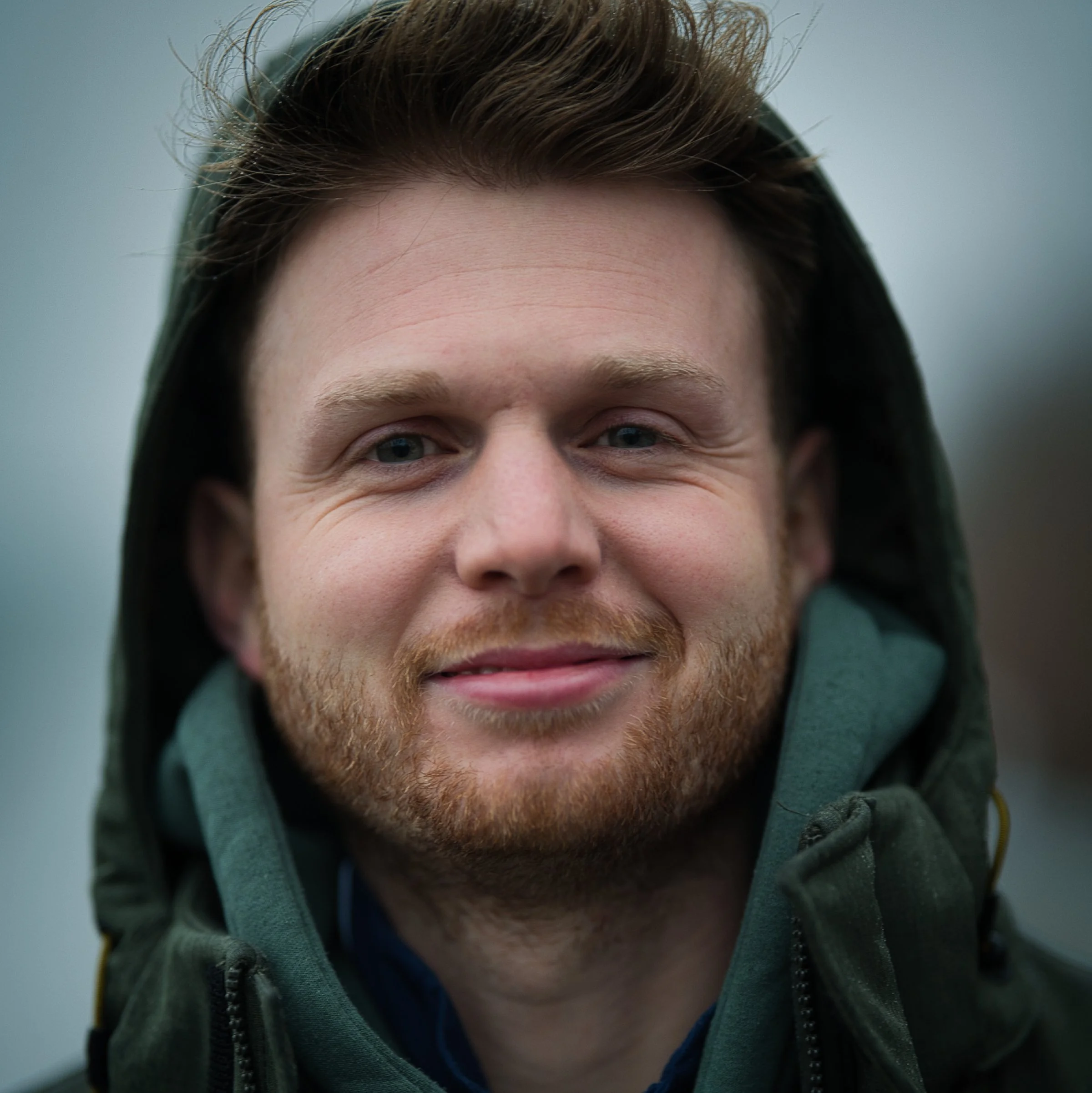 Nahaufnahme des Gesichts eines jungen Mannes mit braunen Haaren, blauen Augen, Bart und Grübchen, trägt einen grünen Hoodie und eine grünen Reißverschluss-Jacke