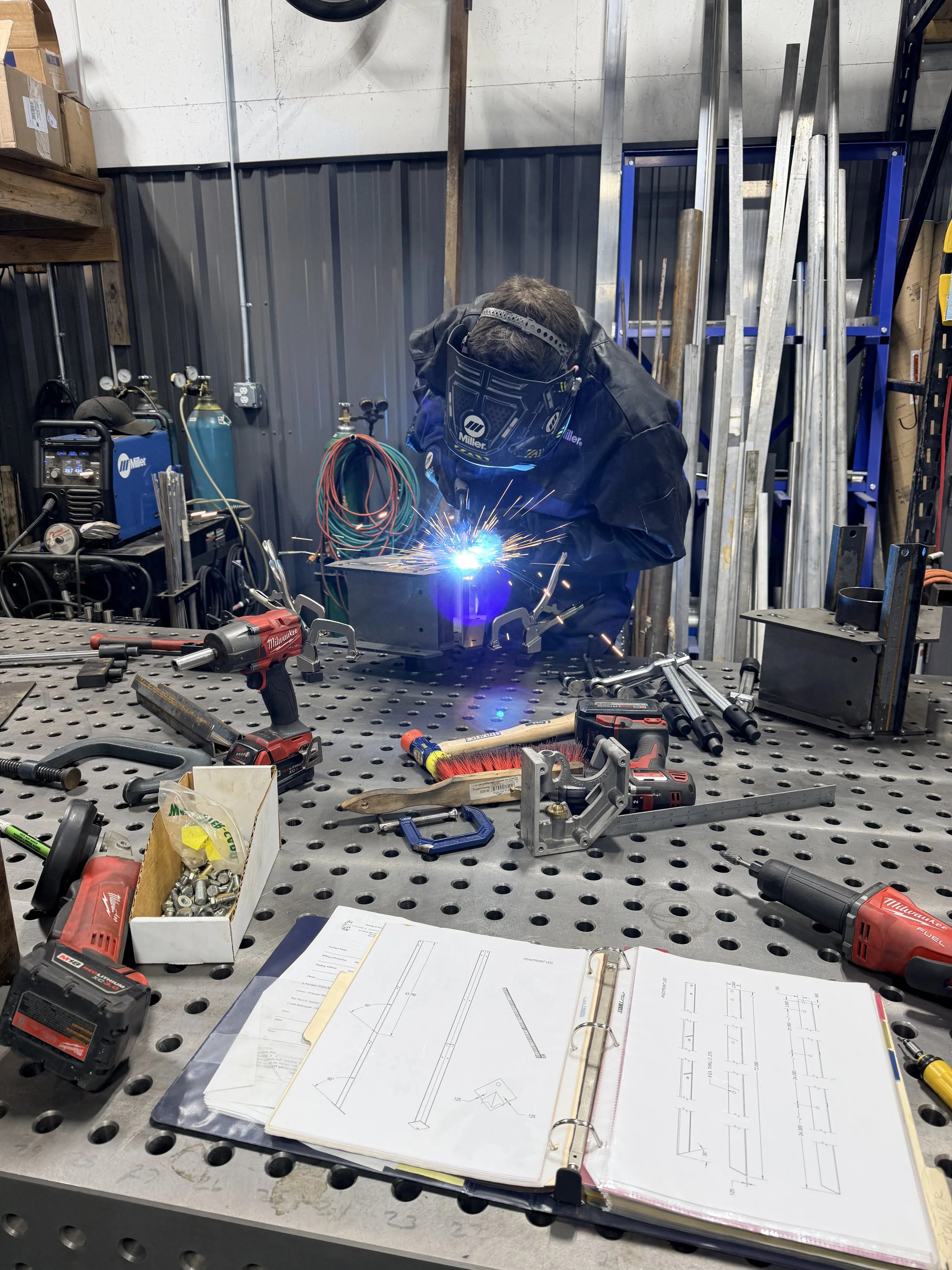 A person welding at a workbench in a metalworking shop surrounded by tools, safety equipment, and architectural plans.