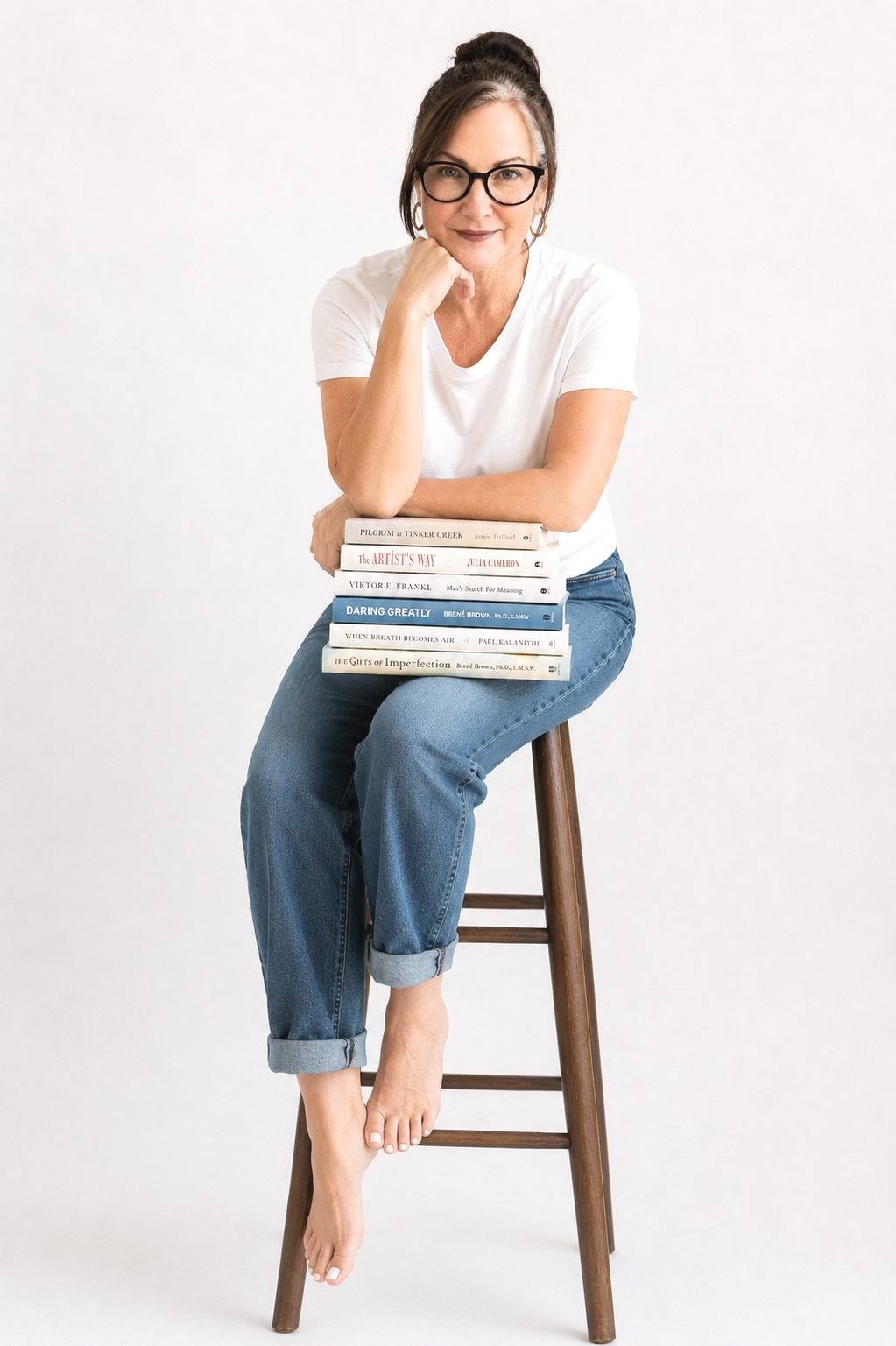 A woman with glasses and dark hair pulled back in a bun, wearing a white t-shirt and jeans, sitting barefoot on a wooden stool, resting her chin on her hand, with a stack of books on her lap.