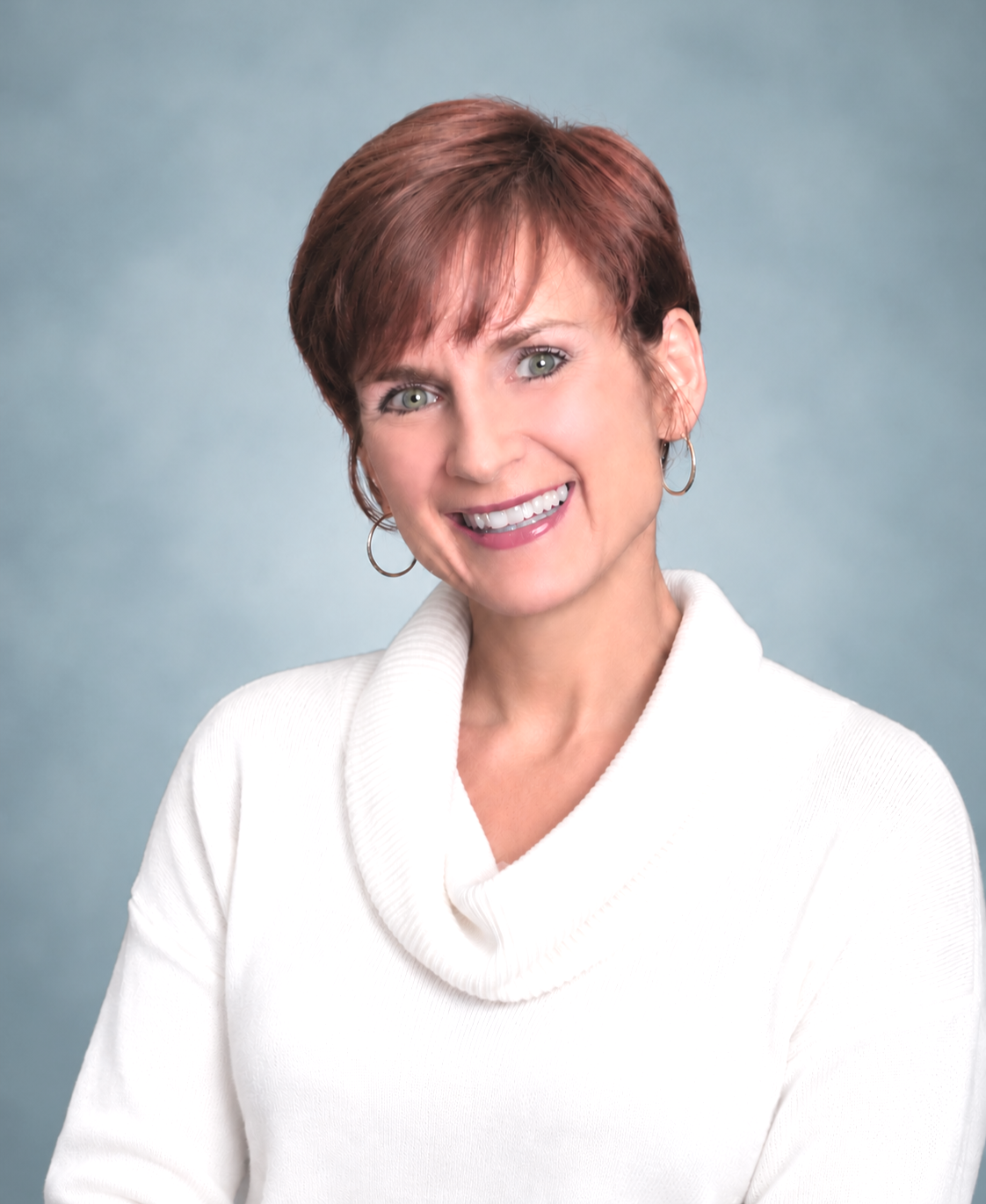 A smiling woman with short red hair wearing a white sweater and hoop earrings, against a light blue background.