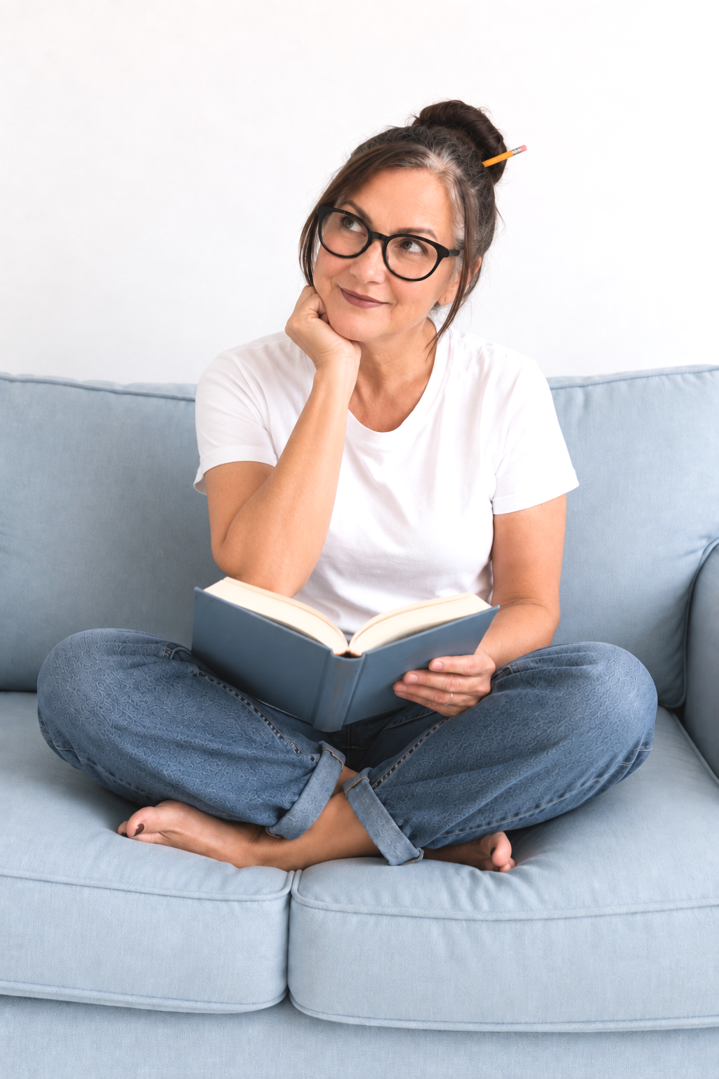 A woman with glasses, gray hair in a bun with a pencil, sitting cross-legged on a couch, holding an open book and thinking.