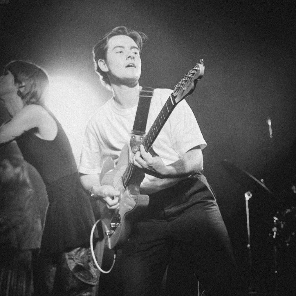 A black and white photo of a young man playing an electric guitar on stage, with a woman in the background wearing glasses and a sleeveless top.
