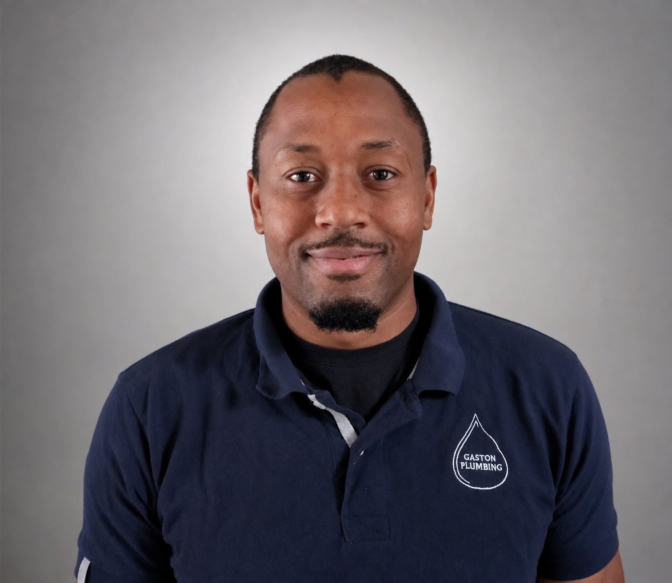 A man with short hair and a small goatee wearing a navy blue polo shirt with a Gaston Plumbing logo, smiling, against a gray background.