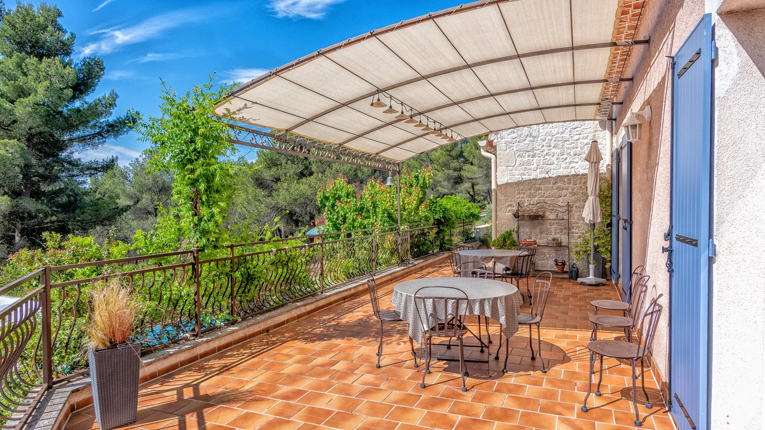 Terrasse ensoleillée avec tables rondes et chaises, protégée par une pergola, entourée de végétation verte et d'un ciel bleu.