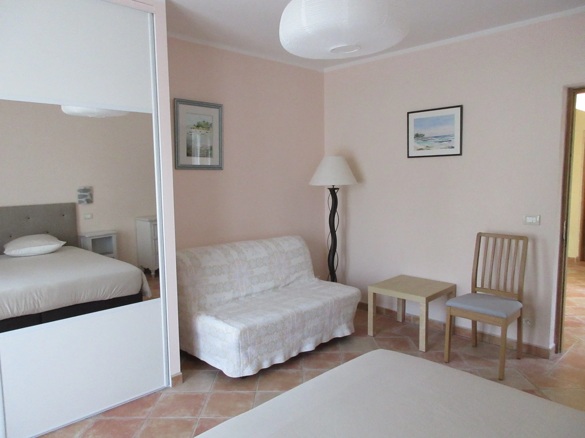 Intérieur d'une chambre avec un lit, un canapé, une lampe, deux tableaux et une table en bois, avec un sol en carreaux.