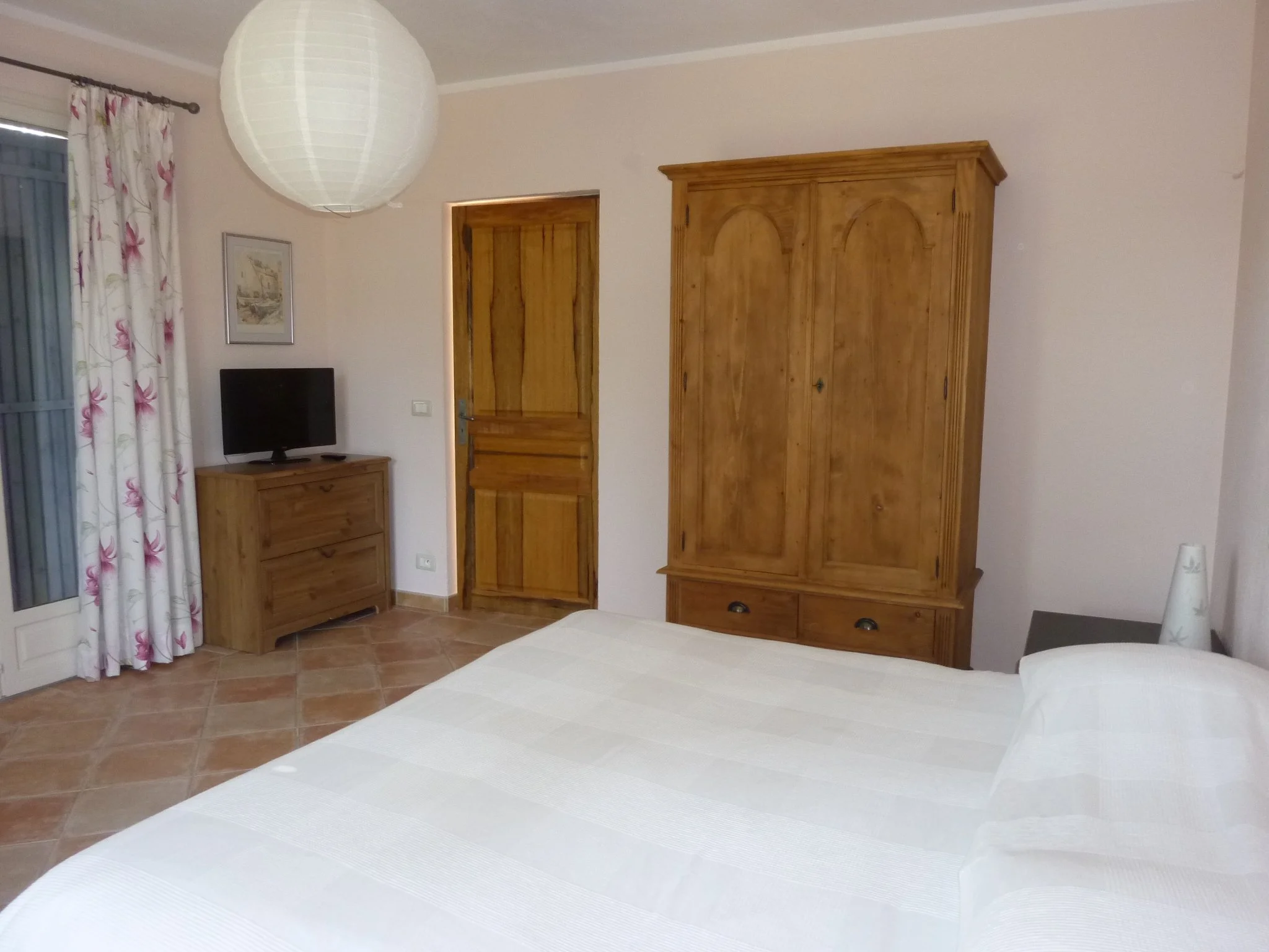 Photo d'une chambre avec un lit, une armoire en bois, une télévision sur une petite commode, une lampe et une fenêtre avec des rideaux à fleurs