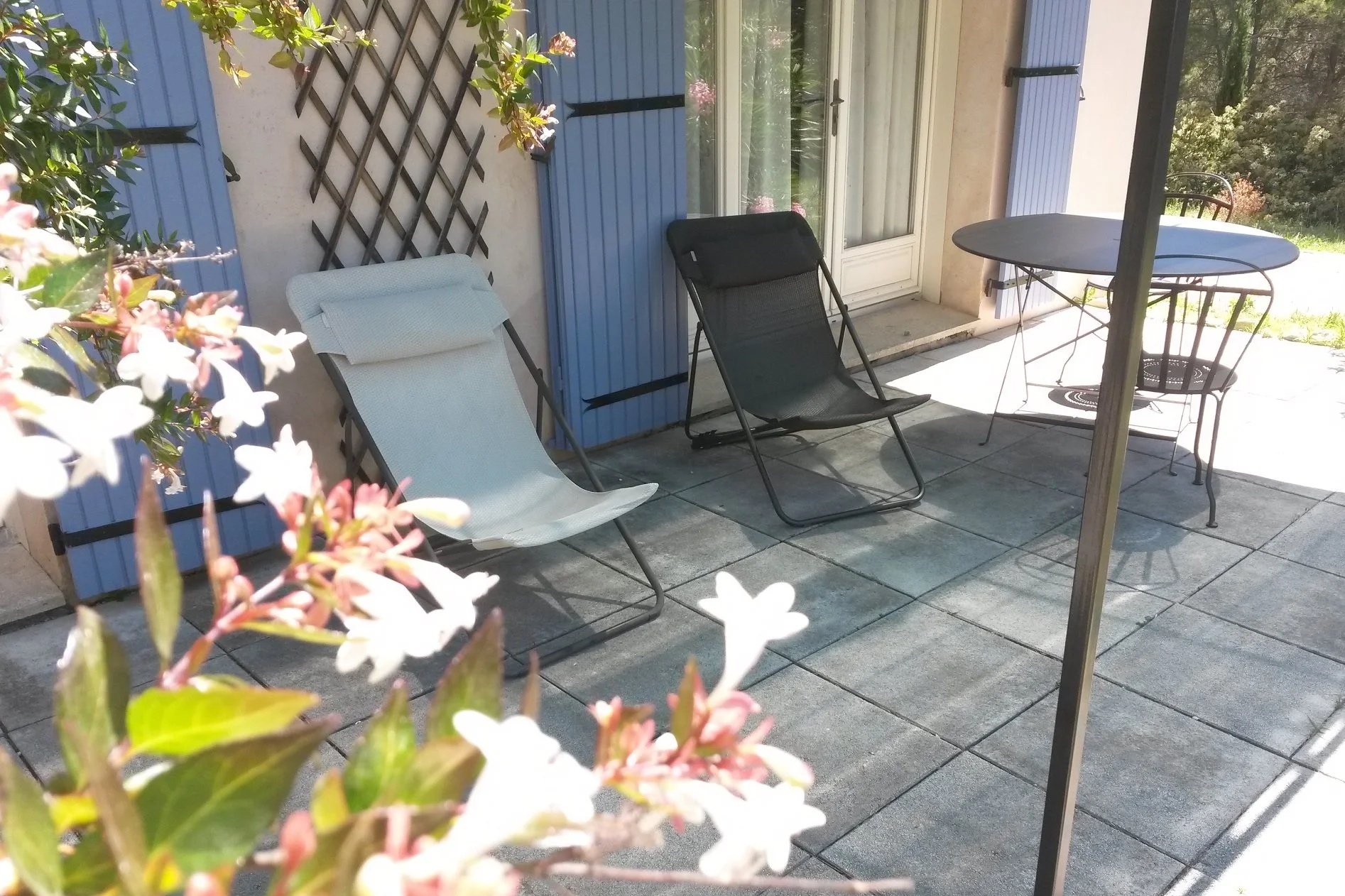 Terrasse ensoleillée avec transats, une petite table ronde en metal, une porte-fenêtre avec volets bleus, et des fleurs en premier plan.