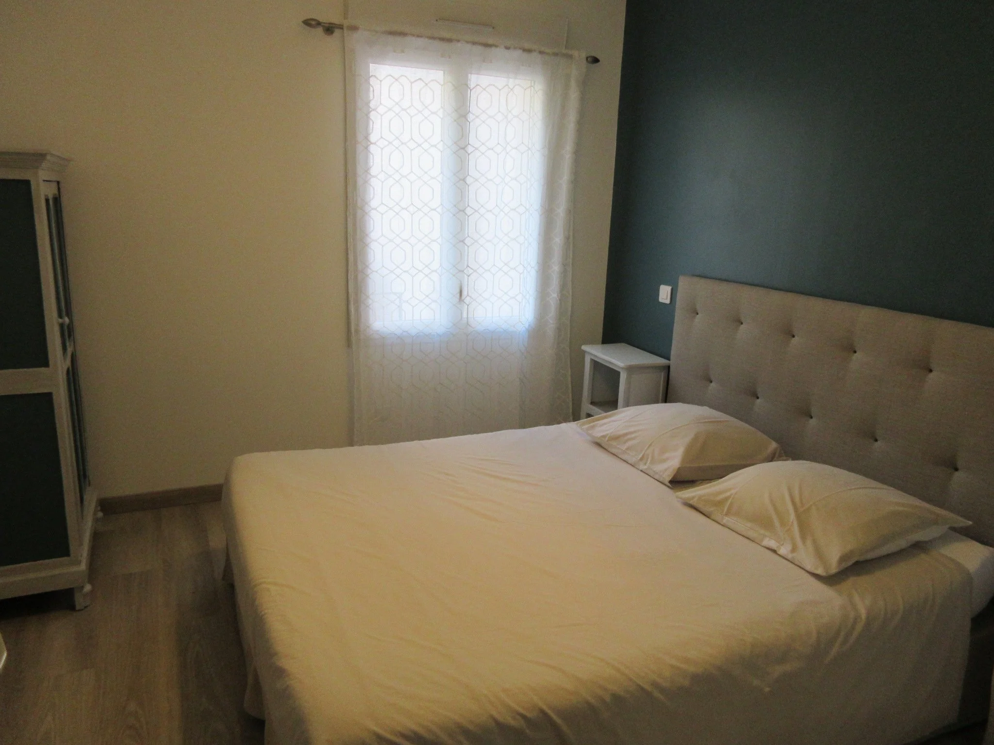 Chambre avec deux lits simples avec draps beige, une tête de lit capitonnée beige, un voilage blanc avec un motif géométrique sur la fenêtre, un meuble blanc à côté du lit et une armoire en partie visible à gauche.