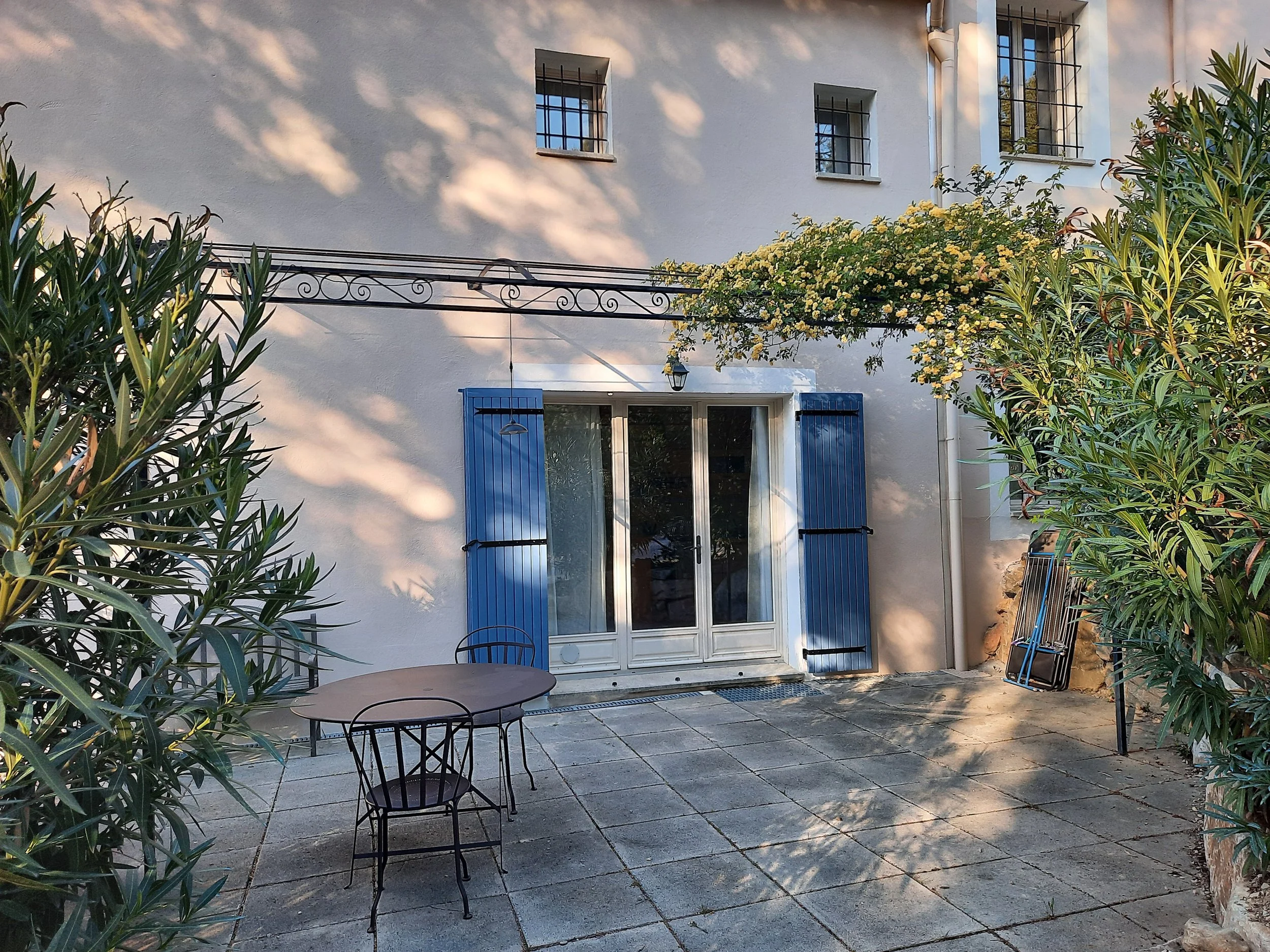 Une terrasse pavée entourée de végétation, avec une maison en arrière-plan. La maison a des fenêtres avec des volets bleus et une porte-fenêtre donnant sur la terrasse. Il y a une table ronde en métal accompagnée de deux chaises.