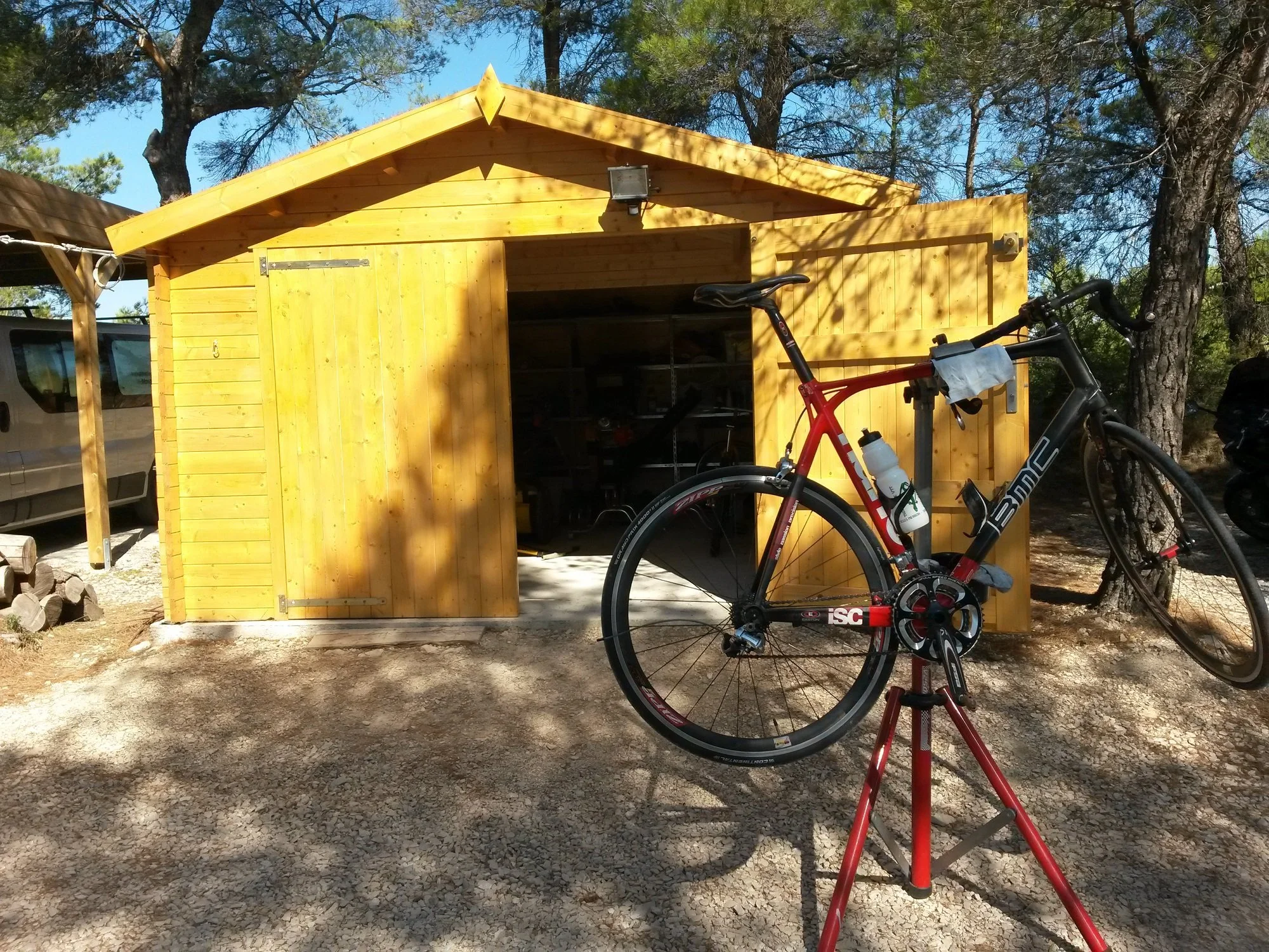 Un abri en bois avec un vélo de route appuyé devant, dans un environnement boisé avec des arbres et un ciel bleu.