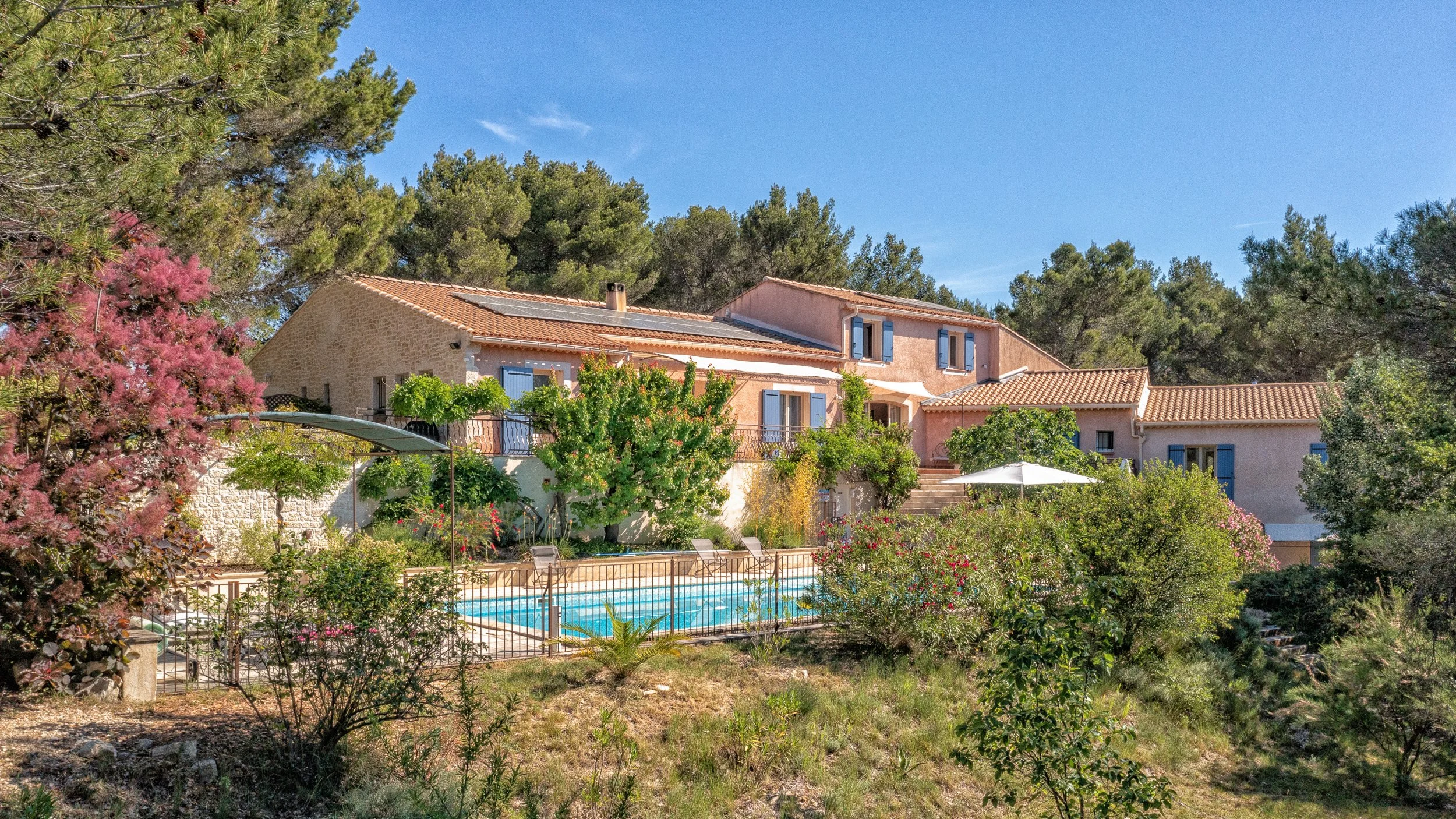 Une maison de style méditerranéen, entourée de nombreux arbres et fleurs, avec une piscine en contrebas
