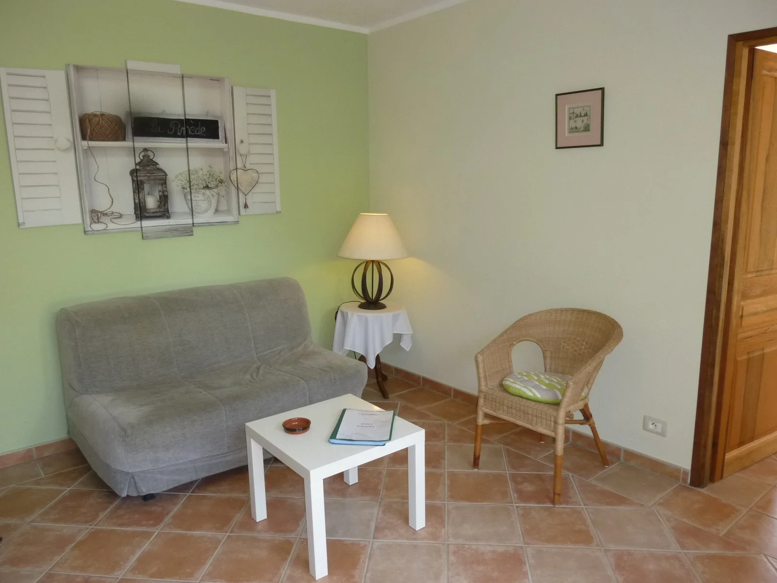Salon avec un canapé gris, une table blanche, une lampe sur une table d'appoint, un fauteuil en rotin avec un coussin rayé vert et blanc, tableau décoratif sur le mur et une porte en bois