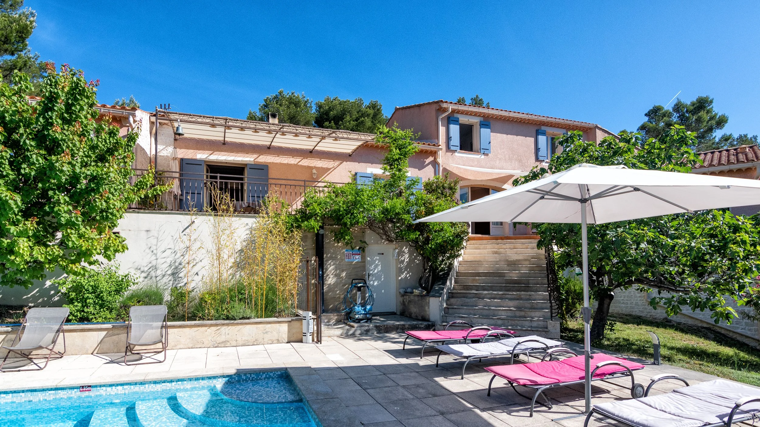 Vue d'une maison avec un jardin et une piscine, avec des chaises longues et une grande parasol. La maison possède une terrasse avec des escaliers et des fenêtres à volets bleus, entourée de verdure et de grands arbres. Ciel clair.