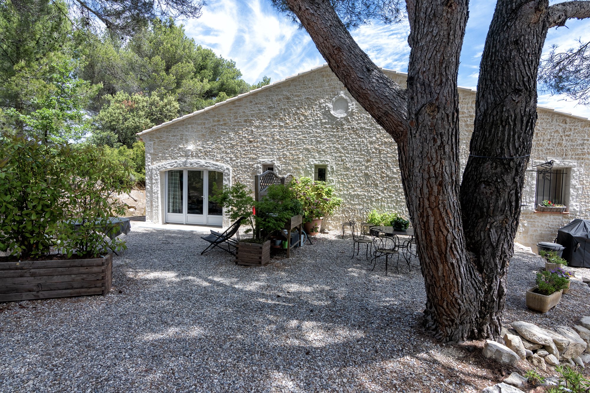 Une maison en pierre, arbres, mobilier de jardin et soleil.