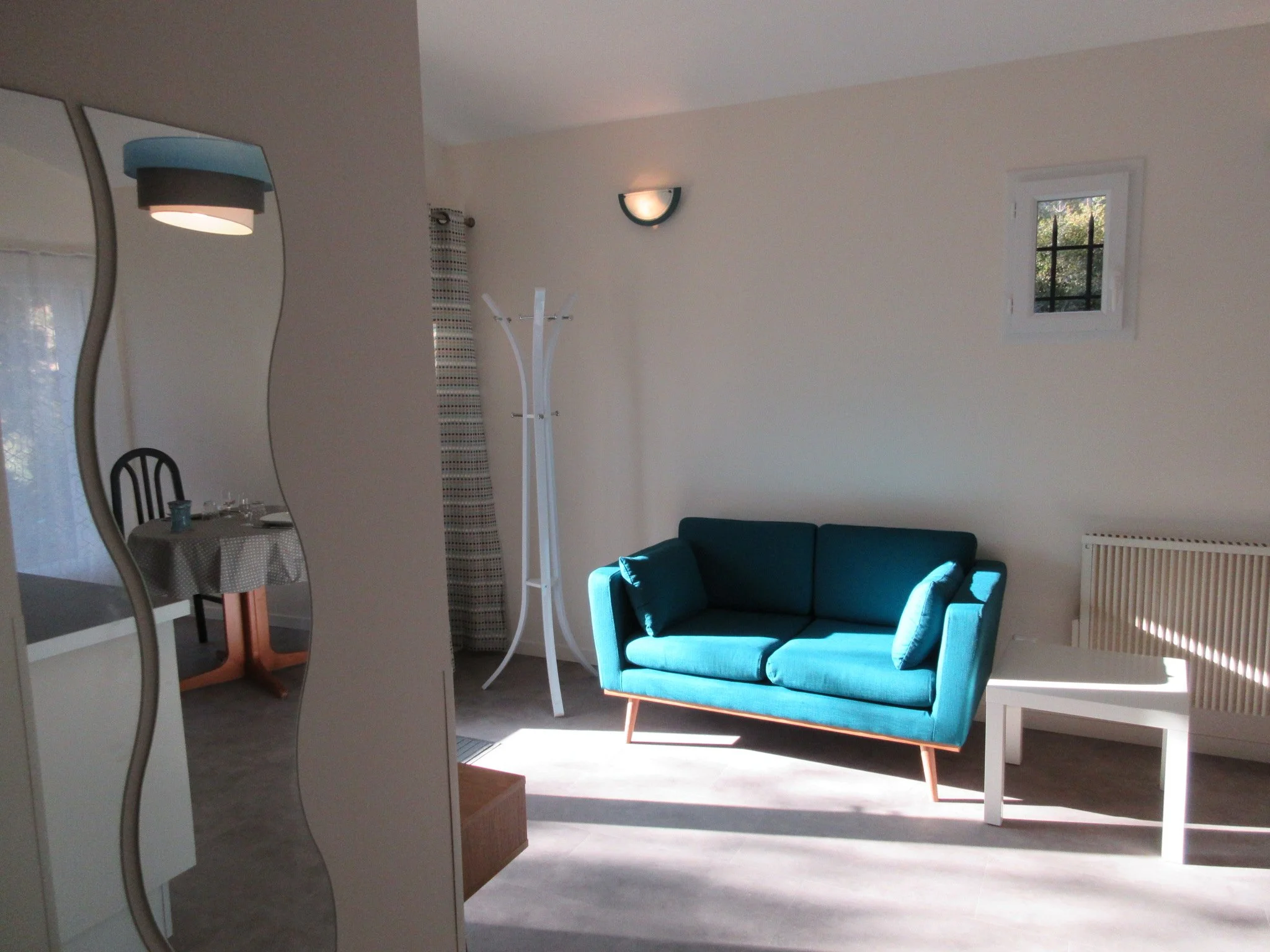 Salon avec un canapé bleu, une lampe murale, un mur blanc, une petite fenêtre, et une chaise noire autour d'une table avec une nappe