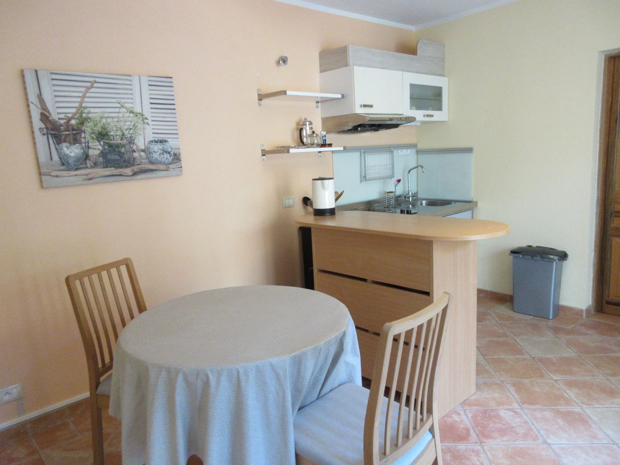 Une cuisine et une salle à manger avec un mur peint en jaune, un tableau représentant des objets de décoration avec des vases, une table ronde avec une nappe blanche, et des chaises en bois. La cuisine a des armoires blanches et un plan de travail en pierre, avec quelques ustensiles et appareils électroménagers visibles.