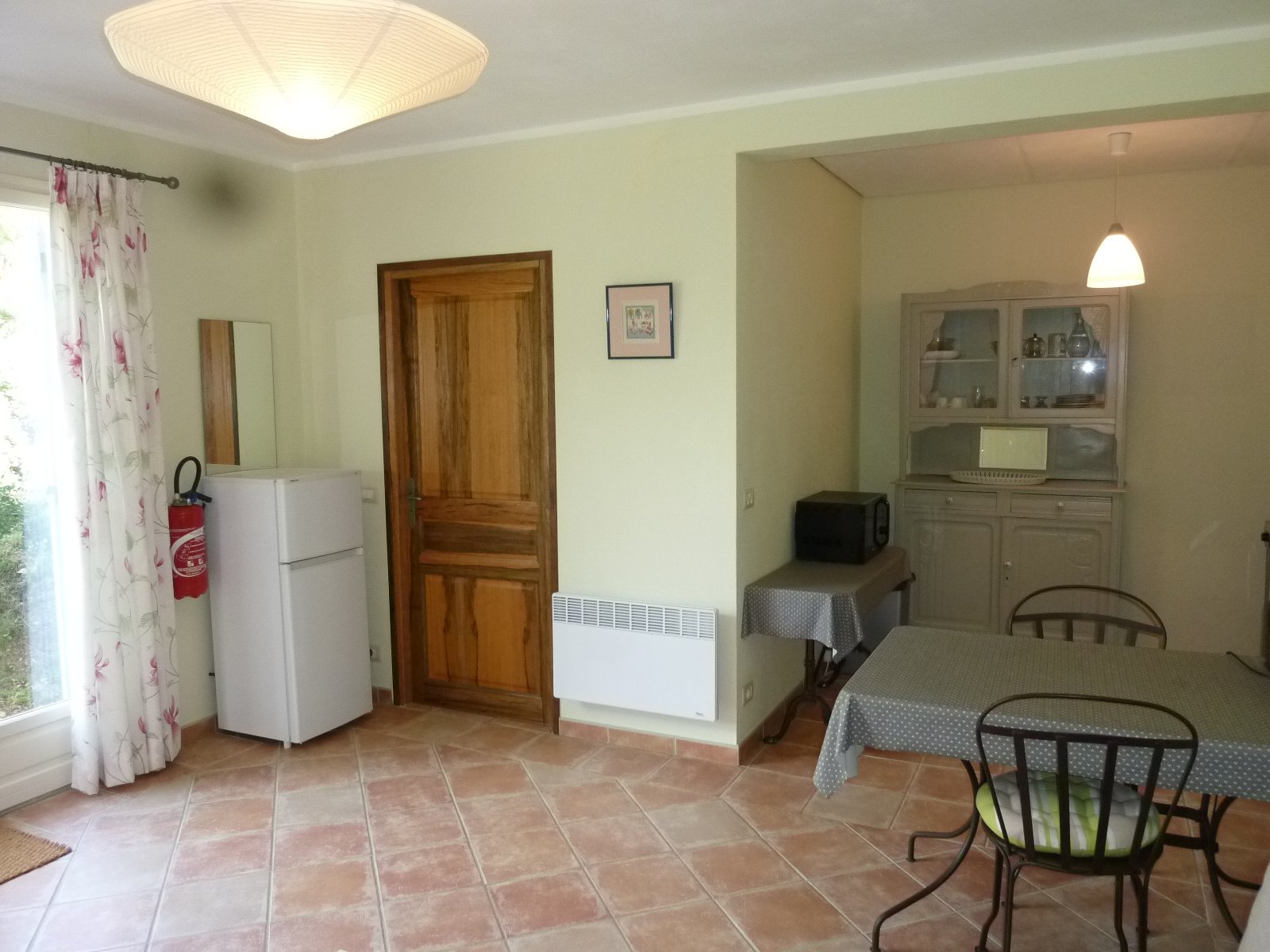 Une cuisine dans une maison avec un réfrigérateur blanc, une porte en bois, une armoire en bois peint en gris, une table recouverte d'une nappe à pois et des chaises en métal. Il y a aussi une fenêtre avec des rideaux à motifs floraux et un radiateur électrique.