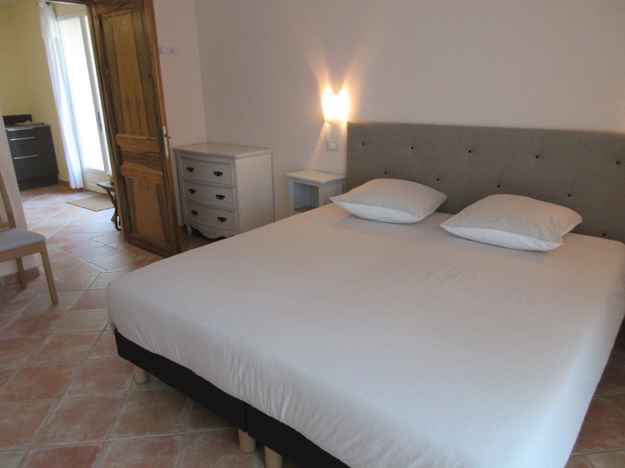 Une chambre avec un lit double à draps blancs, une tête de lit gris, deux tables de chevet blanches, une lampe murale allumée, une porte en bois ouverte et une fenêtre avec rideau blanc laissant entrer la lumière naturelle, un sol en carreaux de terre cuite.