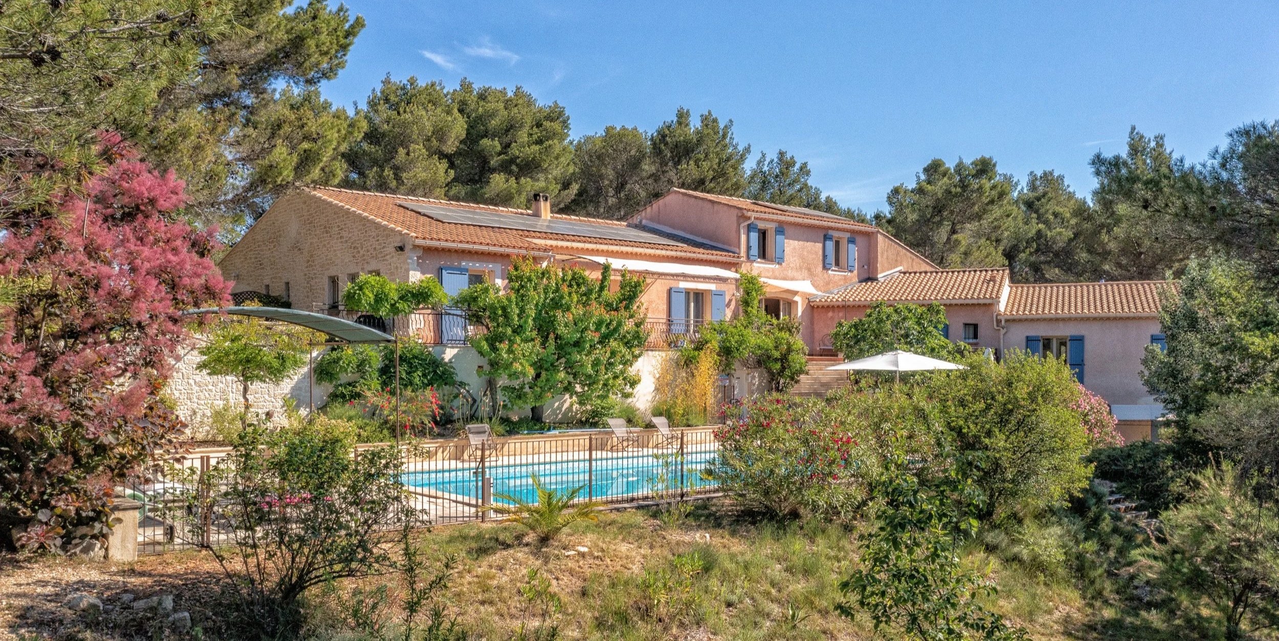 Une maison de style méditerranéen en pierre avec des volets bleus, entourée de végétation luxuriante et un jardin avec une piscine, sous un ciel bleu ensoleillé.