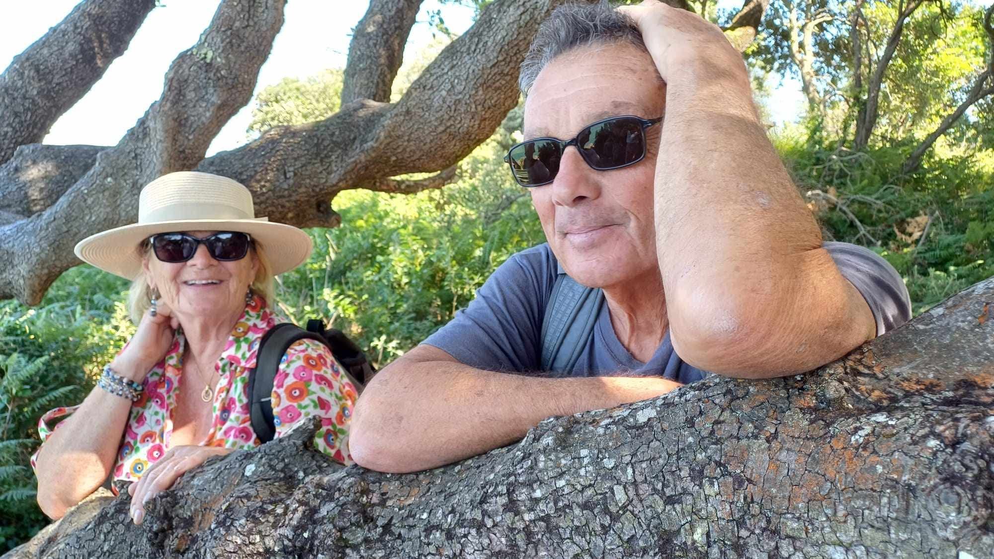 Un homme et une femme profitant d'une sortie en nature, assis sur une branche d'arbre, portant des lunettes de soleil, entourés de verdure et de lumière naturelle.