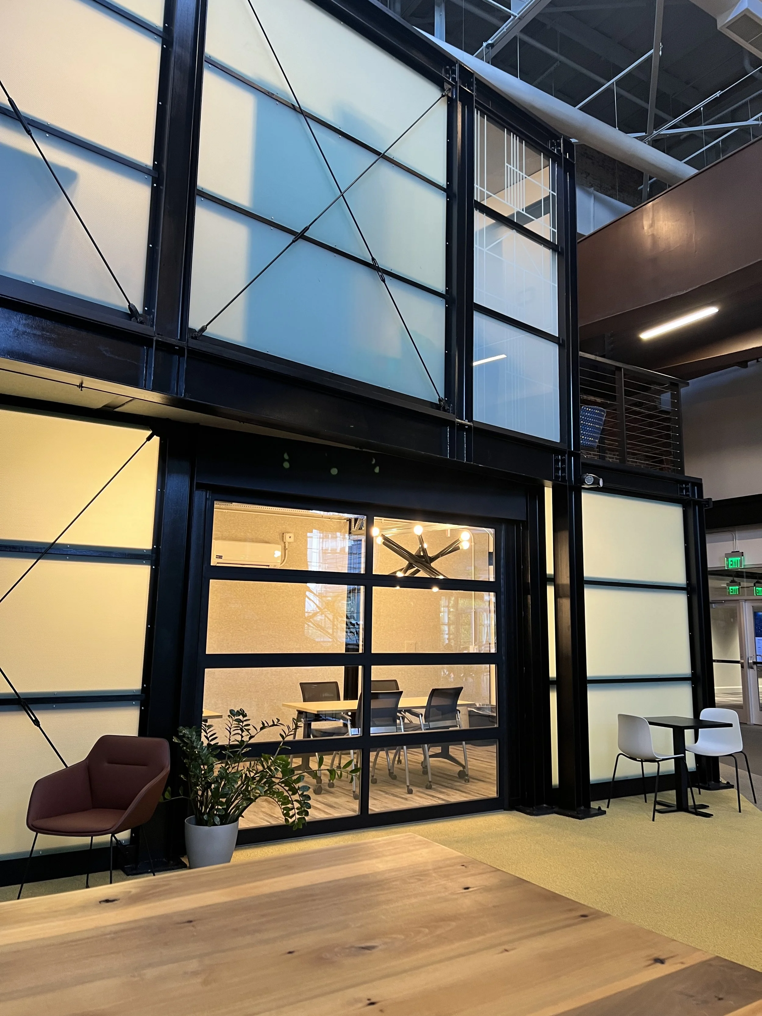 Modern office space with glass partition walls, seating area with chairs and a table, indoor plants, and ceiling lights, in a contemporary building.