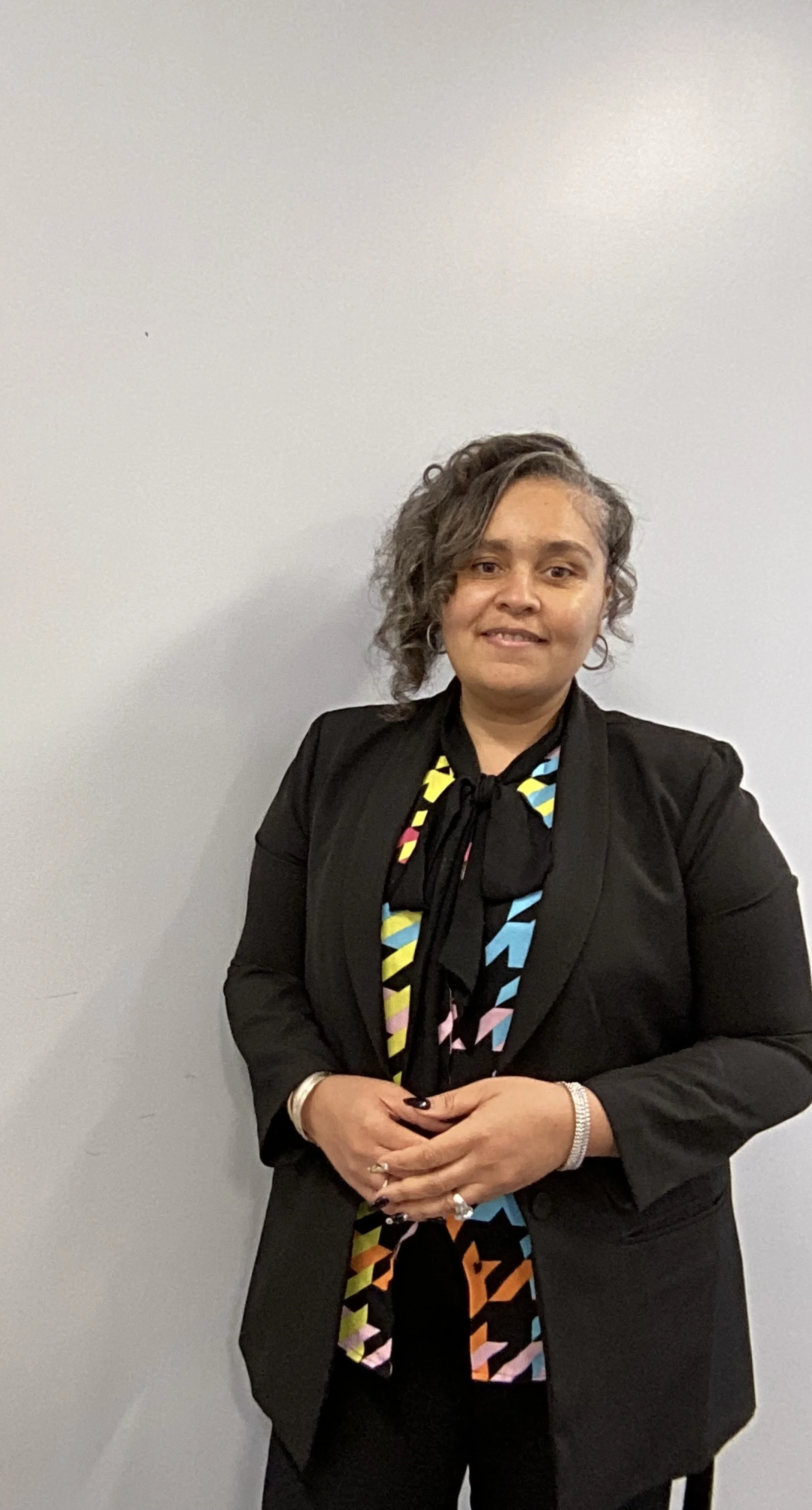 A woman with curly hair wearing a black blazer and a colorful patterned blouse, standing against a plain gray wall.