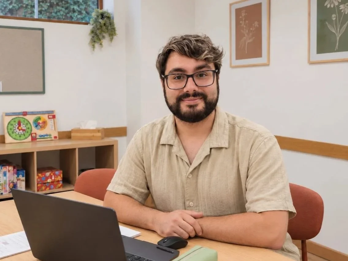 Un hombre con gafas y barba, sentado en una mesa con una laptop, en una oficina con decoraciones de arte y un estante con juegos y libros.