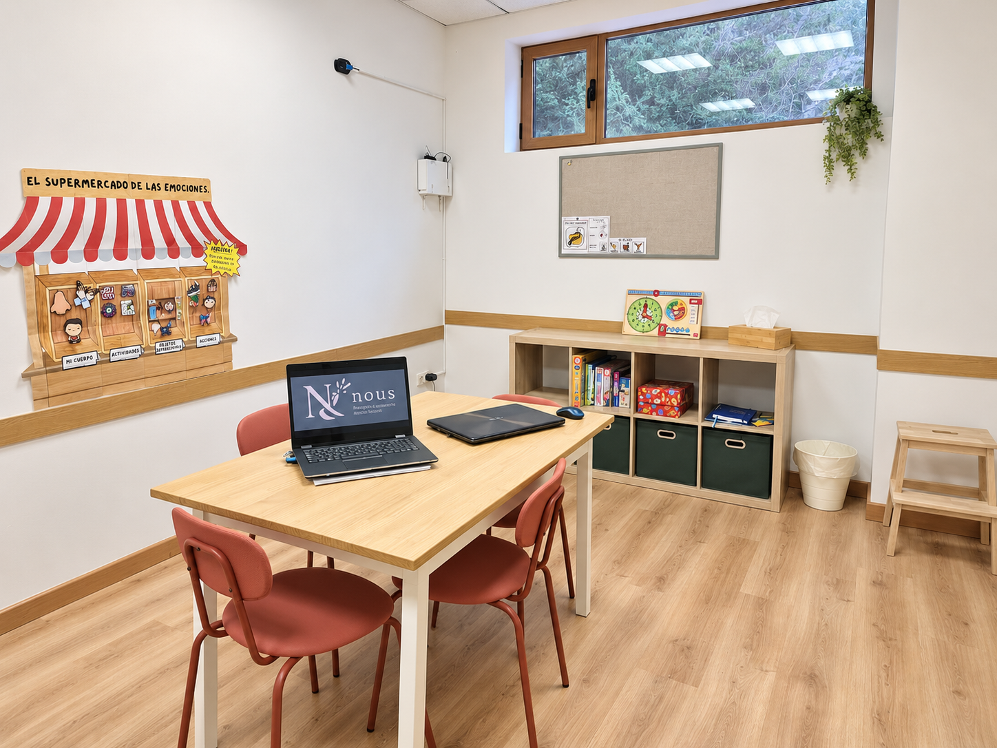 Una oficina pequeña con mesa de madera, tres sillas rosas, una laptop y un mouse. En la pared izquierda, cartel de madera con estantes y figuras decorativas. En la pared de fondo, estantería con libros y cajas, decoraciones infantiles. Ventana grande con vista a árboles. Piso de madera y paredes blancas.
