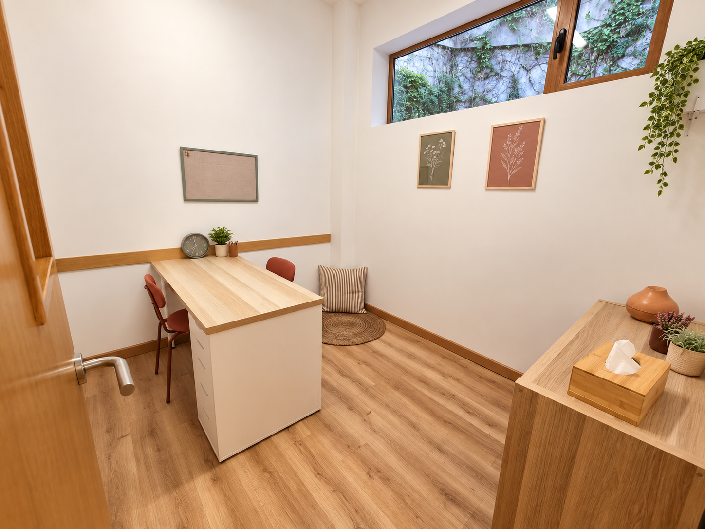 Una oficina pequeña con escritorio de madera, una silla y decoraciones sencillas en las paredes, con una ventana que da a un muro cubierto de plantas.