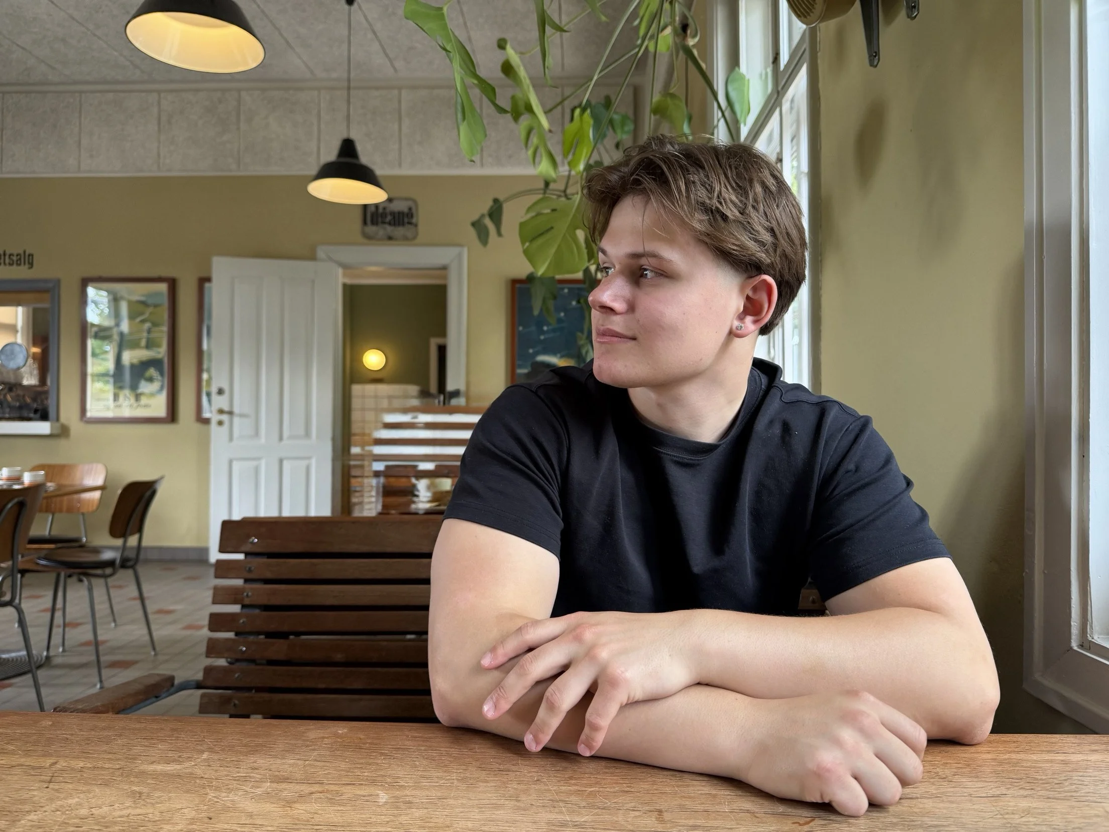 En ung mand sidder ved et træbord i en café eller restaurant, og ser ud gennem et åbent vindue med et afslappet udtryk.