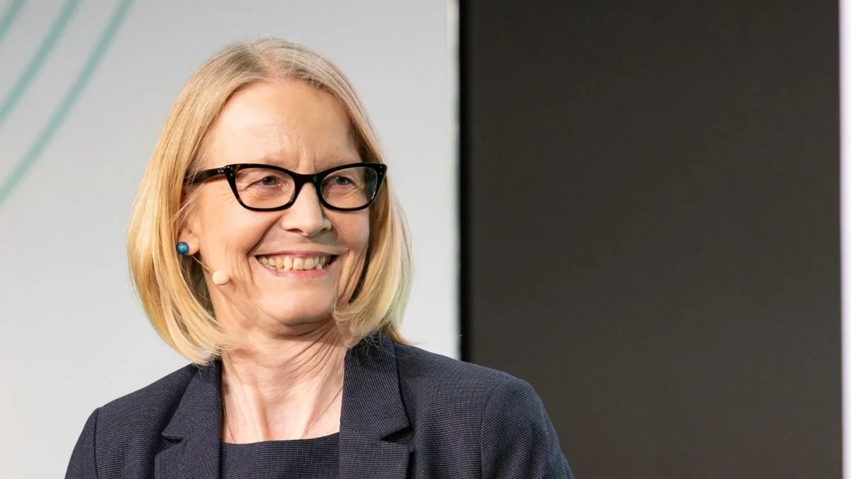 A smiling woman with blonde hair, wearing glasses, a microphone headset, and a dark blazer.