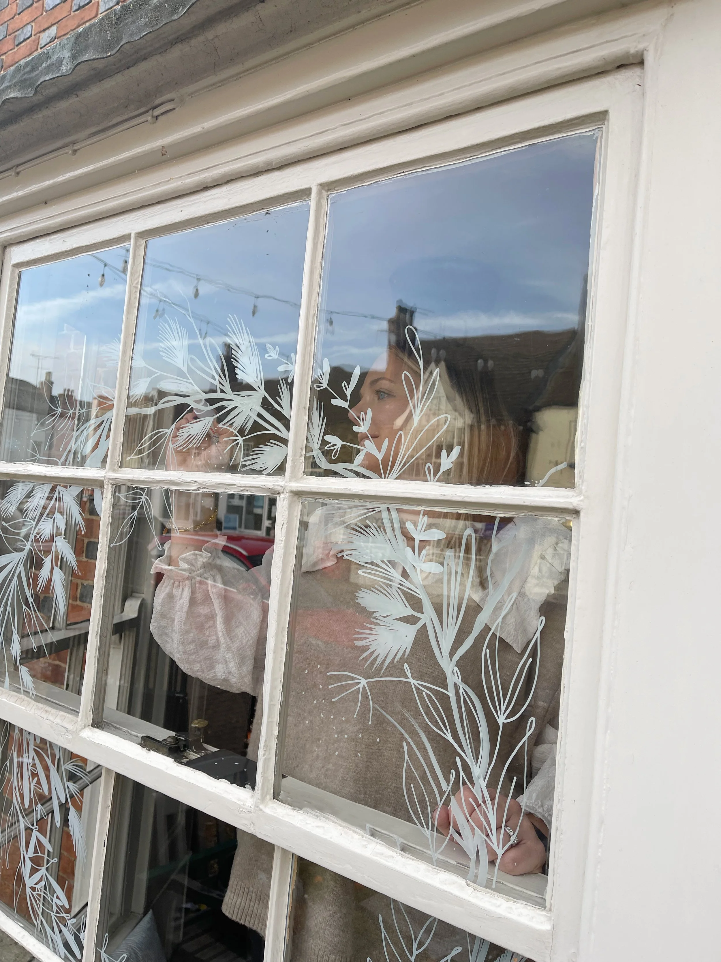 A person decorating or painting a window with white floral designs, viewed from outside.