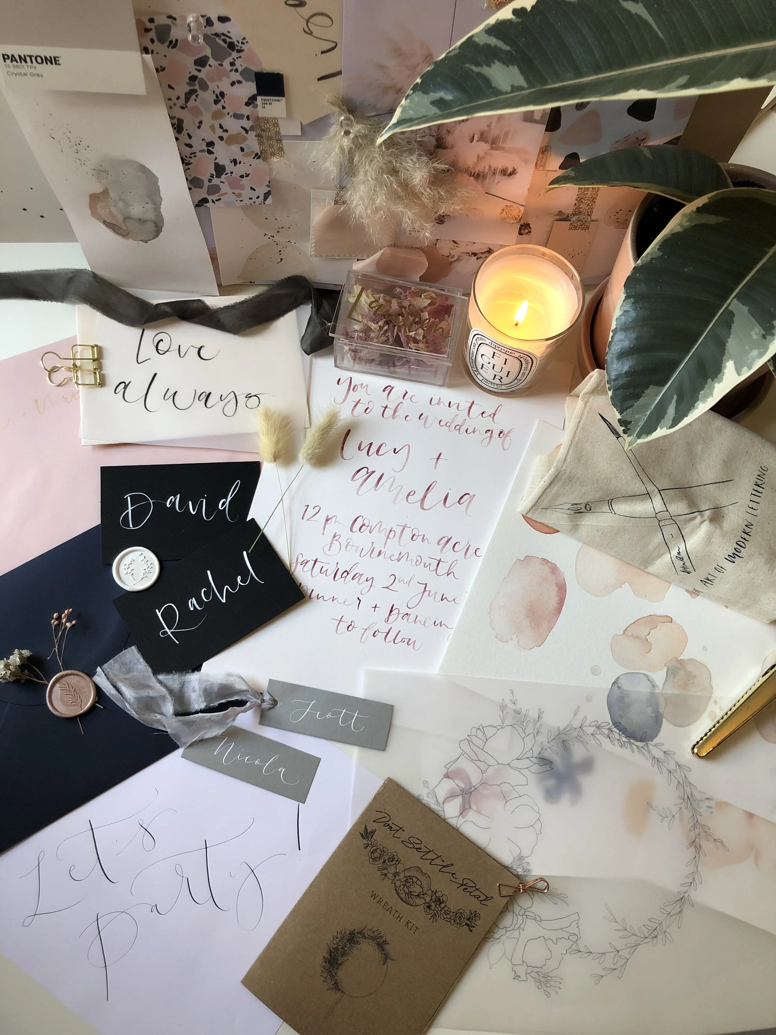 A flat lay of wedding planning items including handwritten wedding invitations, stationery with calligraphy, a lit candle, potted plant, dried flowers, a wax seal, and various decorative papers and tags.