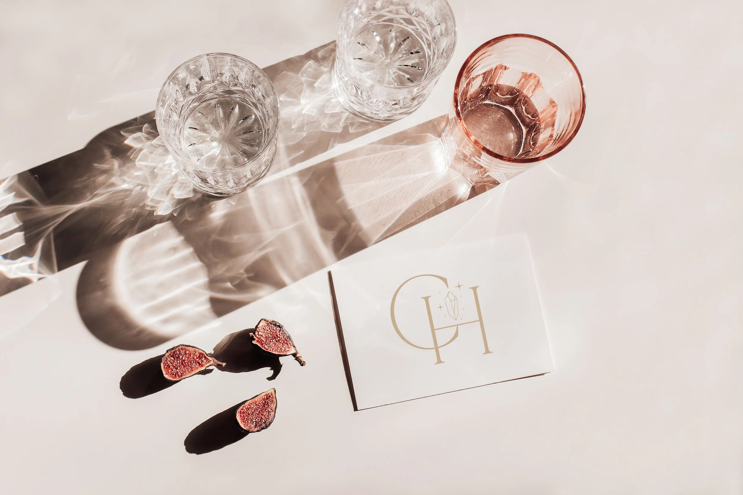 Two clear textured glasses and one pink-tinted glass on a white surface with three slices of dried figs and a white card with gold logo.