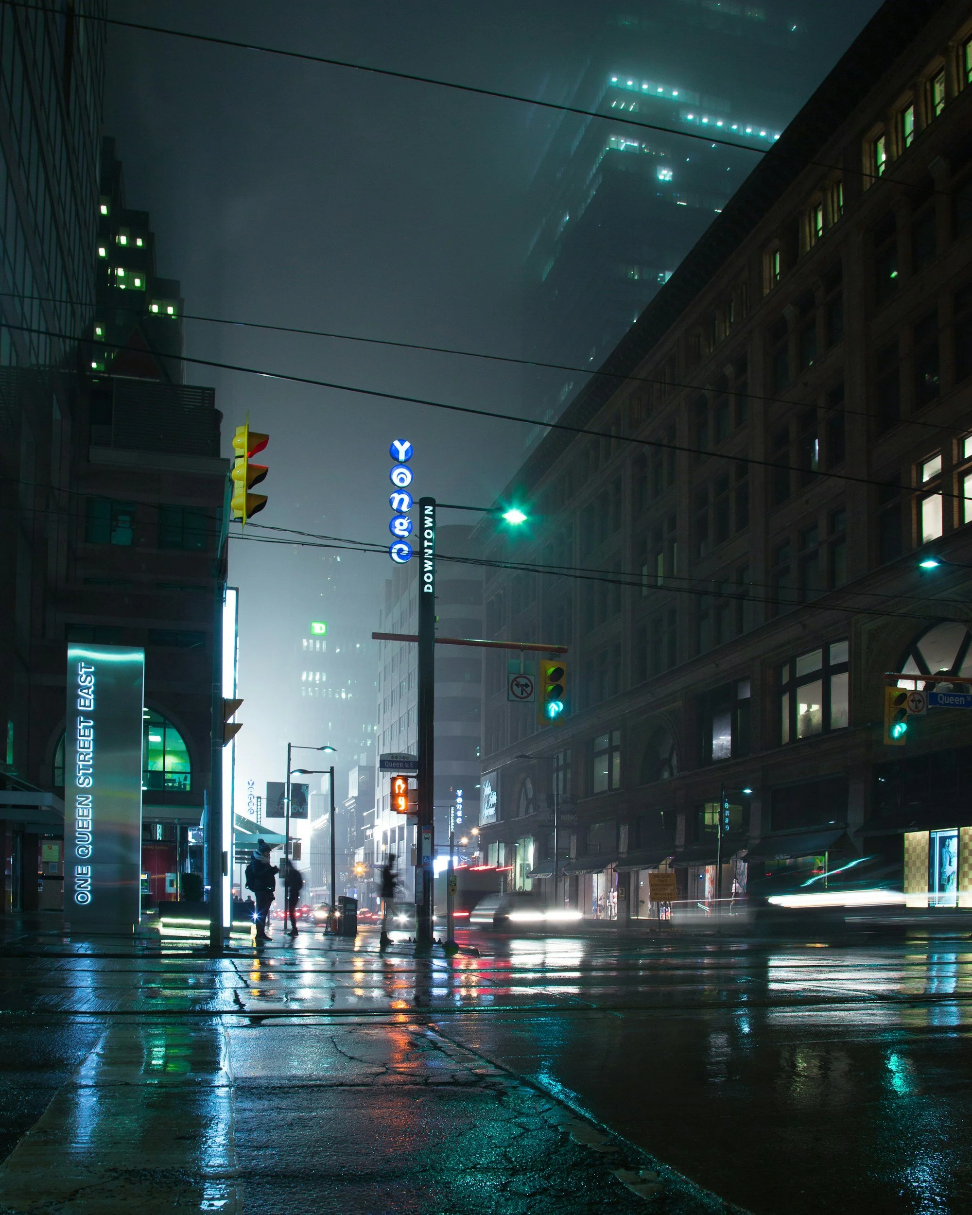 A city street scene at night with wet pavement reflecting lights. Tall buildings with illuminated windows and signage, including a visible sign that says "ONE QUEEN STREET EAST" and a Toronto subway sign reading "DOWNTOWN." Pedestrians are walking, and traffic lights are on, with some car headlights visible.