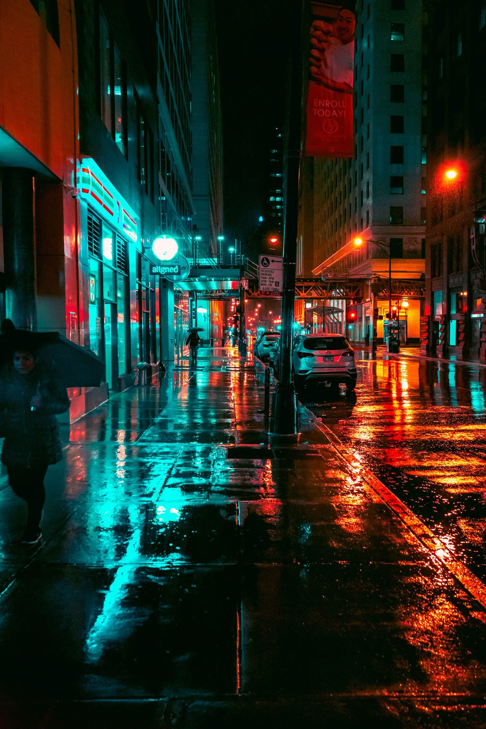 A city street at night after rain, illuminated by colorful neon lights reflecting on wet pavement. Several pedestrians walk with umbrellas, and parked cars line the street.