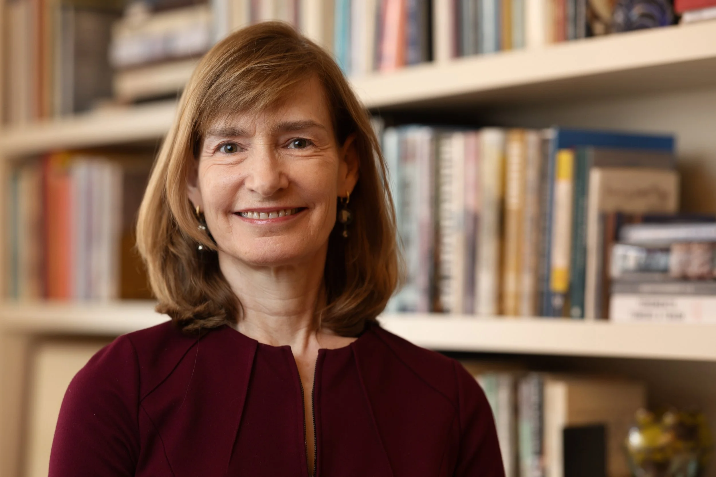 Margi Hofer, a middle-aged woman with shoulder-length brown hair smiling, wearing a burgundy top and earrings, standing in front of a bookshelf filled with books.