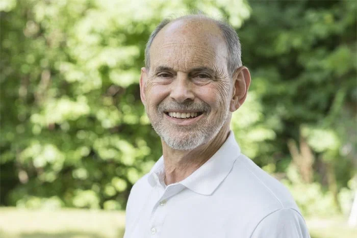 Ward Mintz, a smiling elderly man with a white beard and short hair, wearing a white collared shirt, outdoors with green trees in the background.