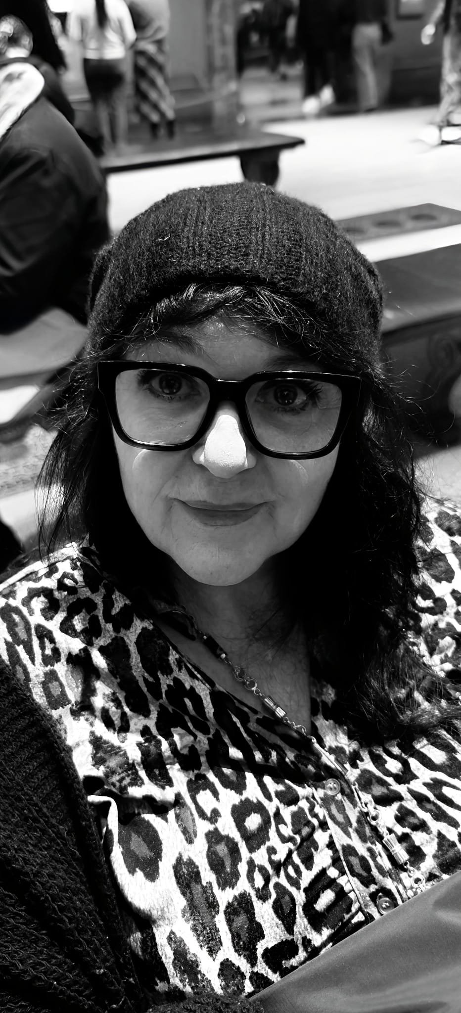 A woman with dark hair, glasses, and a beanie taking a selfie in a public indoor space, wearing a leopard print jacket.