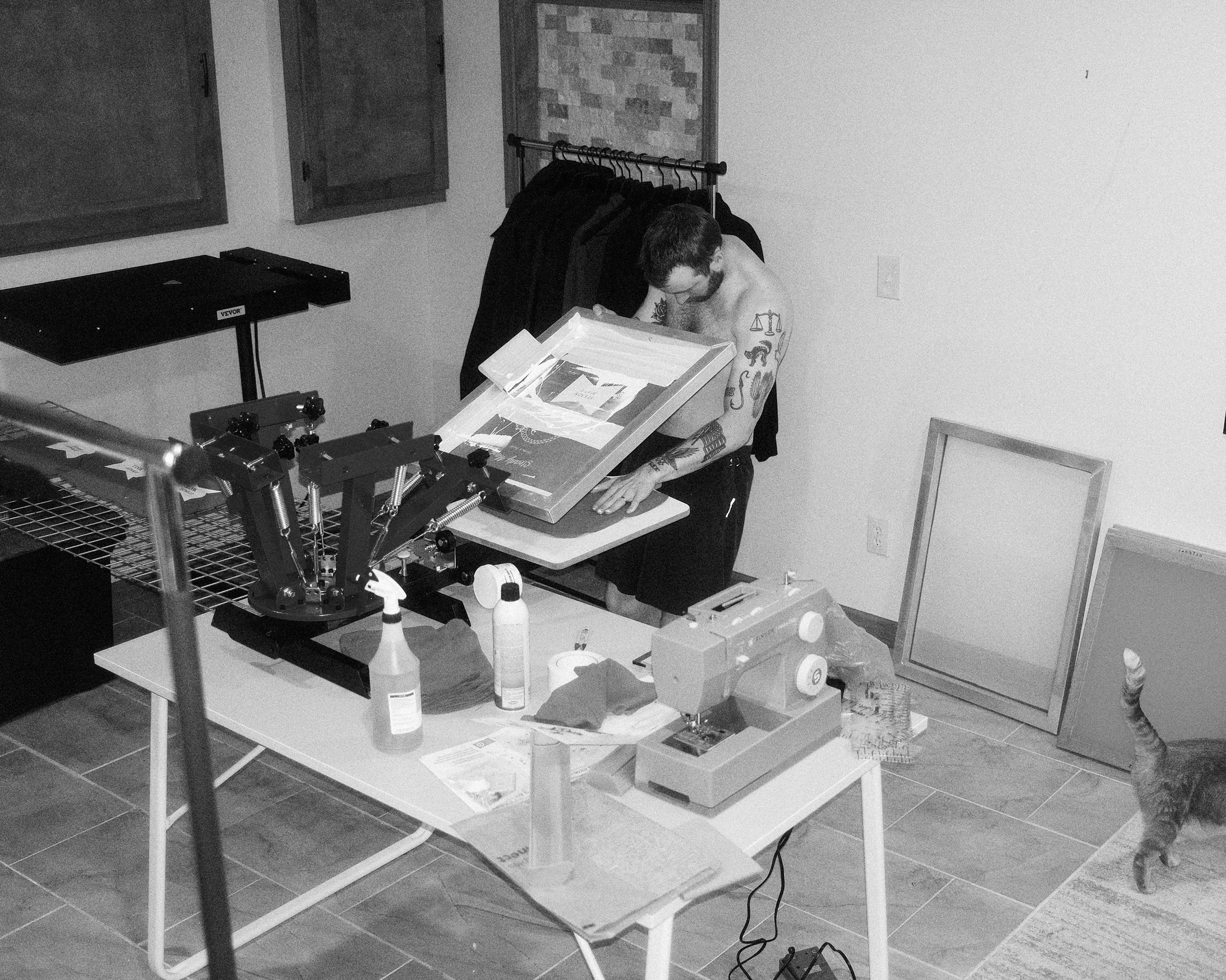 A shirtless man with tattoos on his arms is screen printing merchandise.