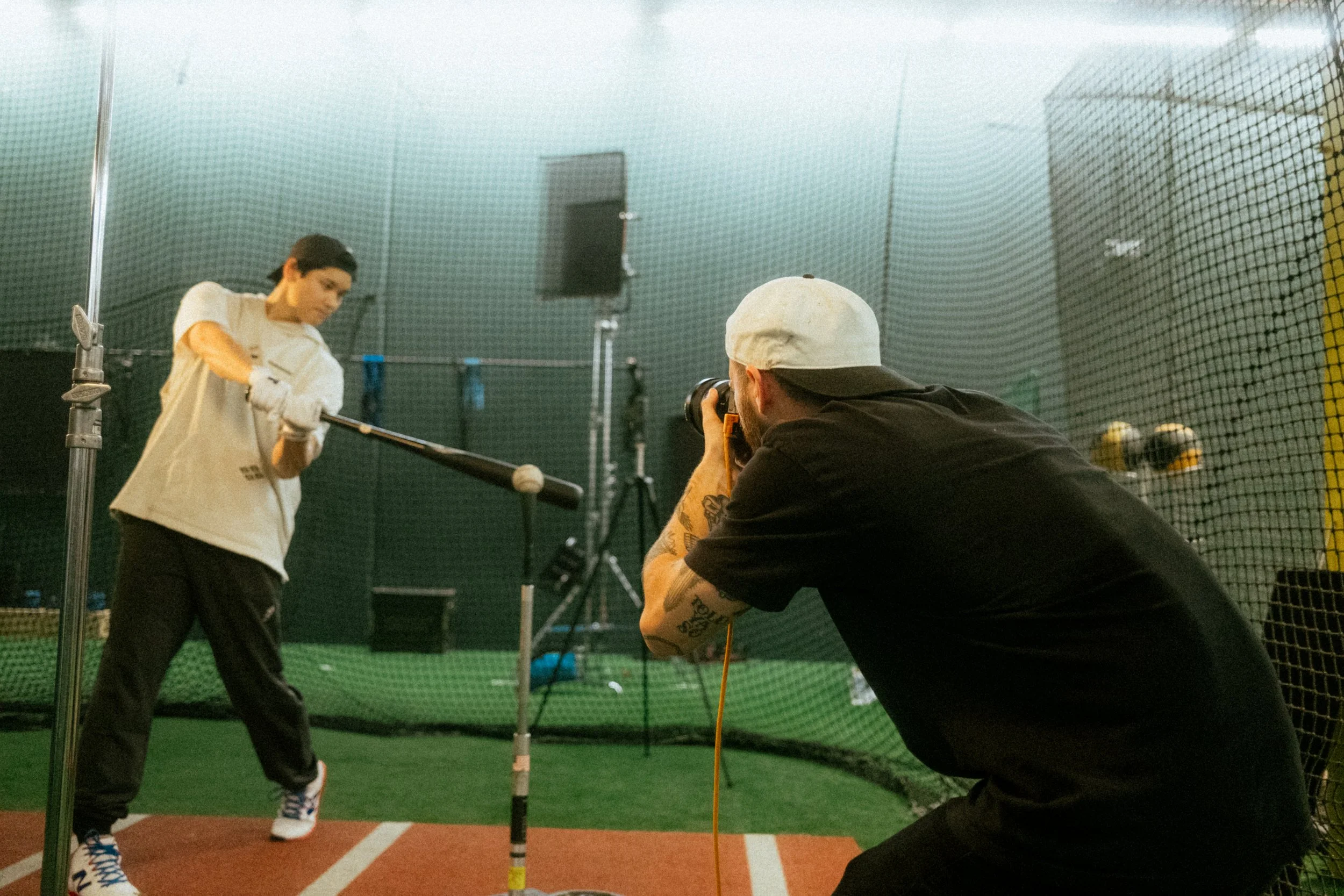 World famous baseball player in a white t-shirt and black pants prepares to hit a baseball with a bat during a photoshoot inside a batting cage. A photographer wearing a black shirt and white cap takes pictures of her through a camera.