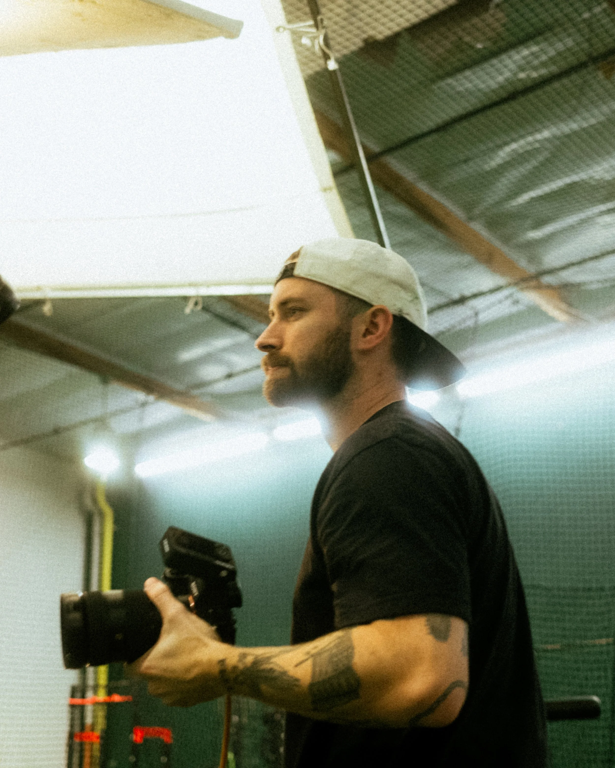 Man with a beard wearing a backward baseball cap and a black t-shirt, holding a camera in a gym. The ceiling has metal panels and exposed beams.