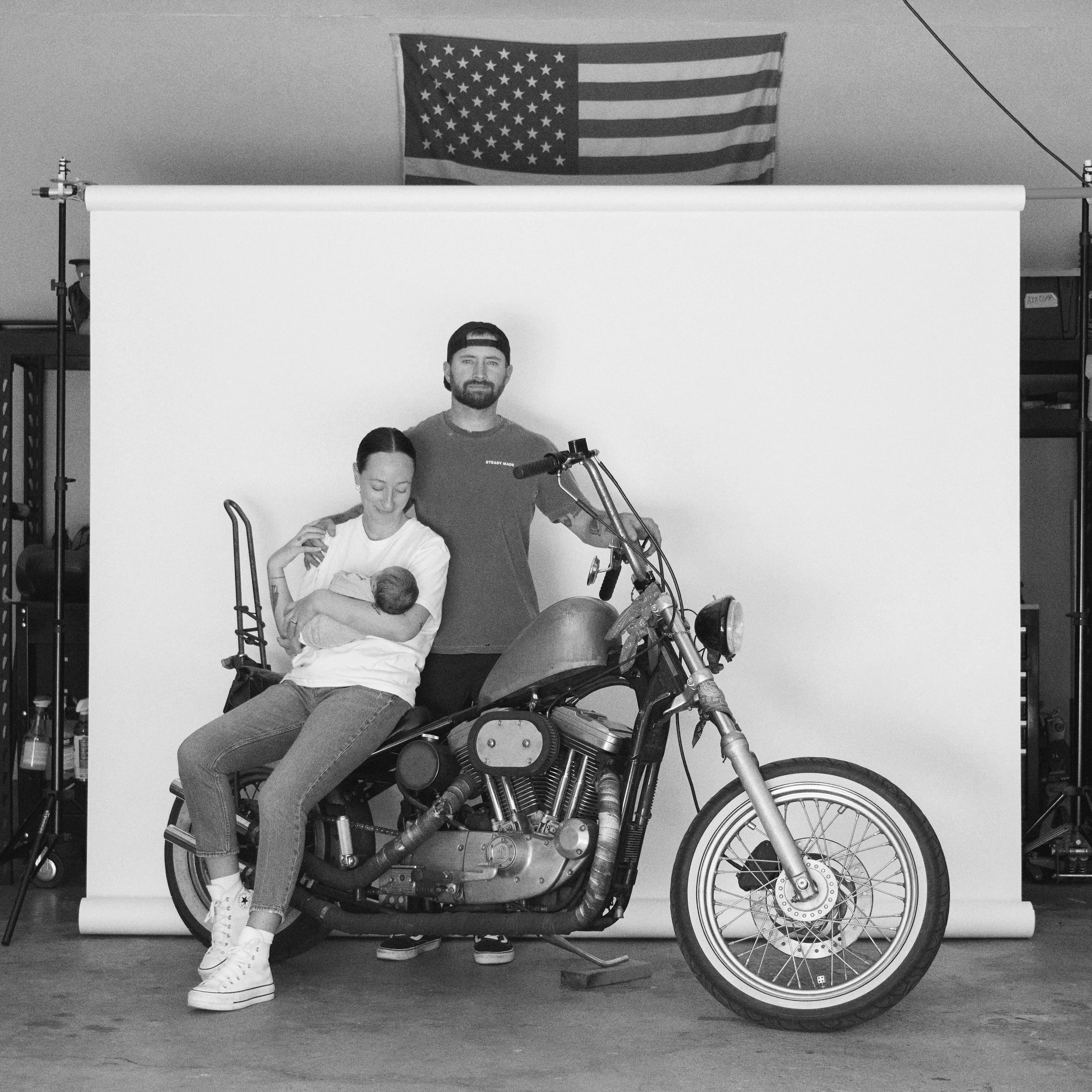 People posing in front of a backdrop with an American flag hanging above, with a motorcycle in the foreground and one person sitting on it, another standing behind, and a child sitting on the person's lap.