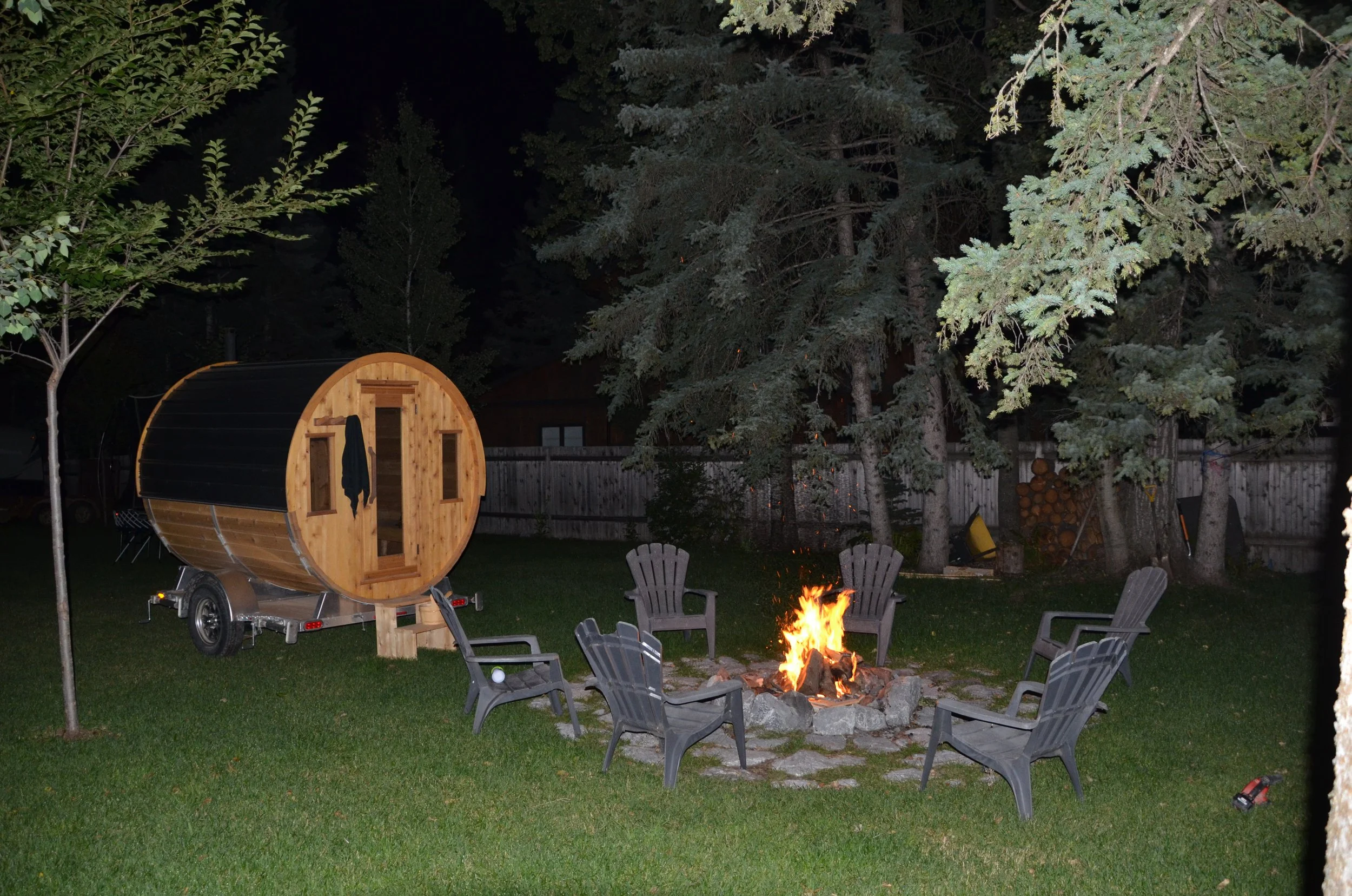 Backyard scene at night with fire pit, multiple patio chairs, a wood burning barrel sauna on wheels, and trees surrounding the area.