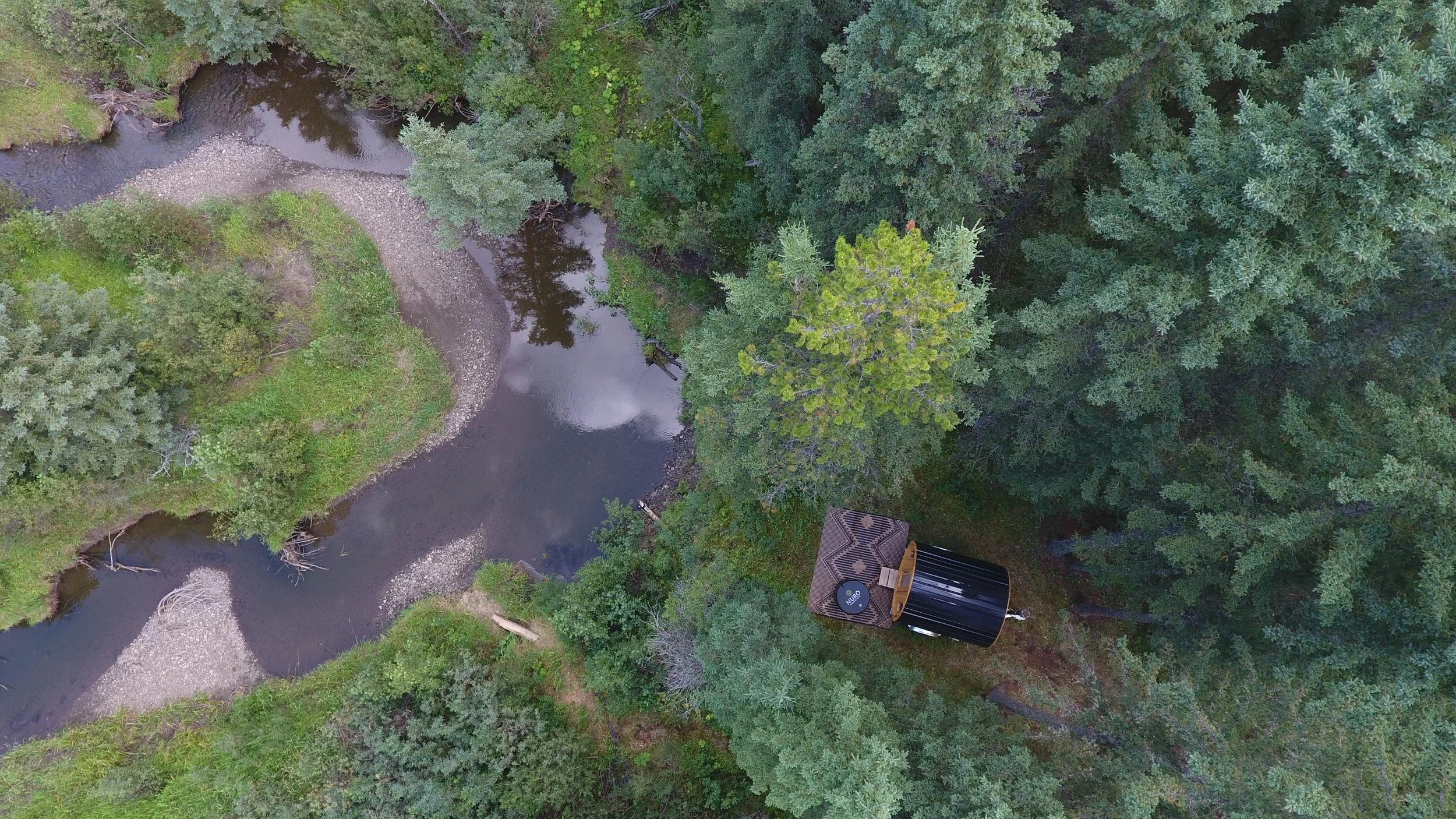 An aerial view of a small creek with dense green trees on both sides, showing a barrel sauna from above overlooking the creek.