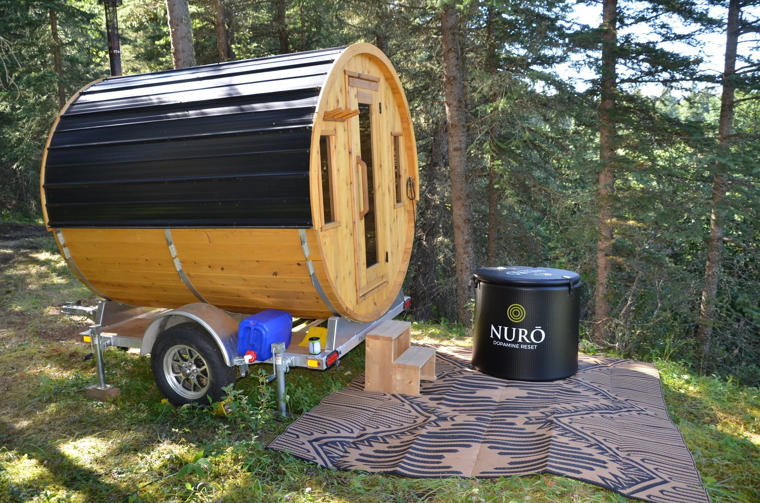 A small wooden sauna with a curved black metal roof on a trailer, set in a forest with pine trees. There is a black cold plunge tub nearby, a small blue water container, and a patterned outdoor rug.